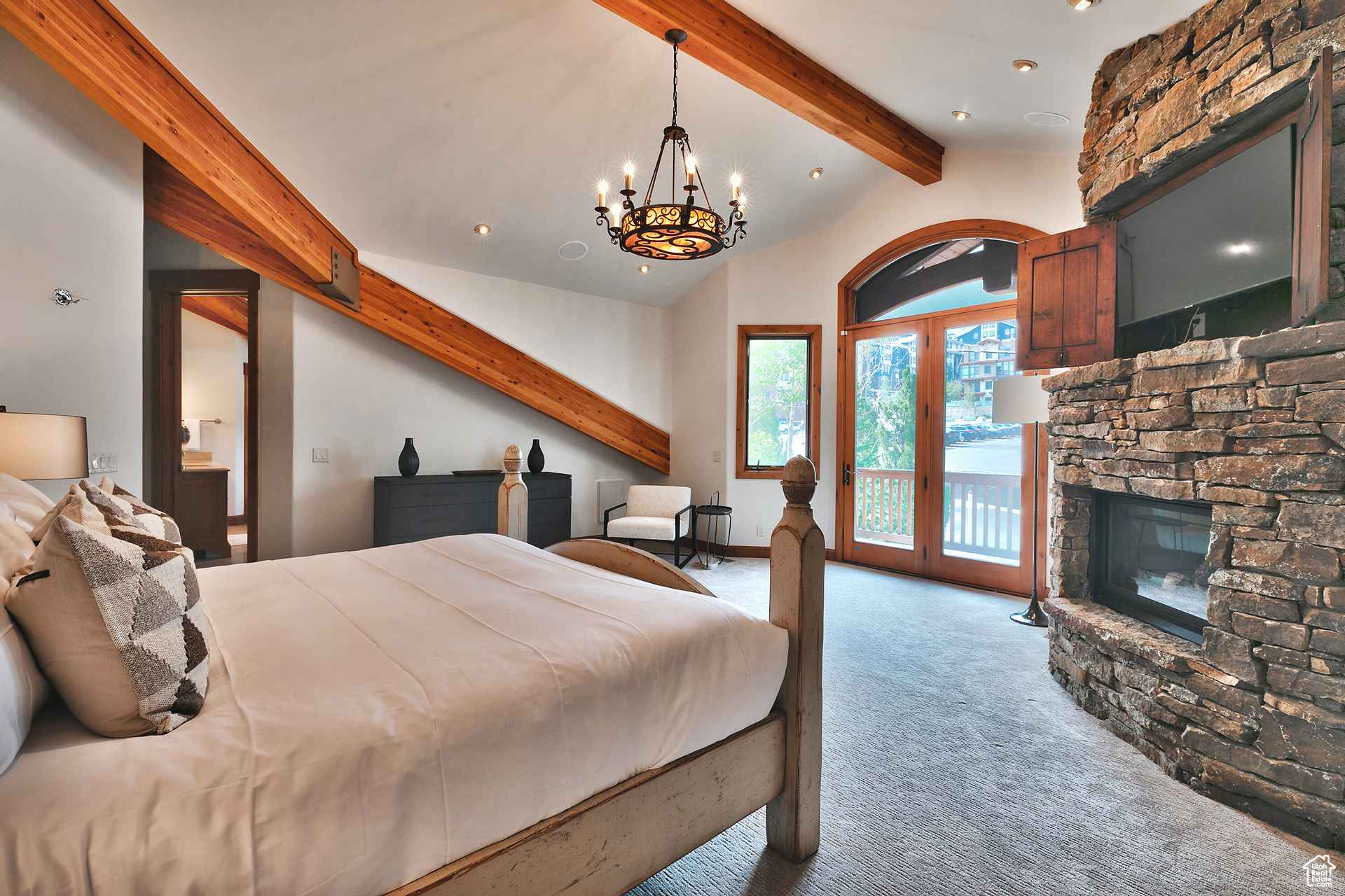 Carpeted bedroom with a stone fireplace, a chandelier, access to exterior, and lofted ceiling with beams