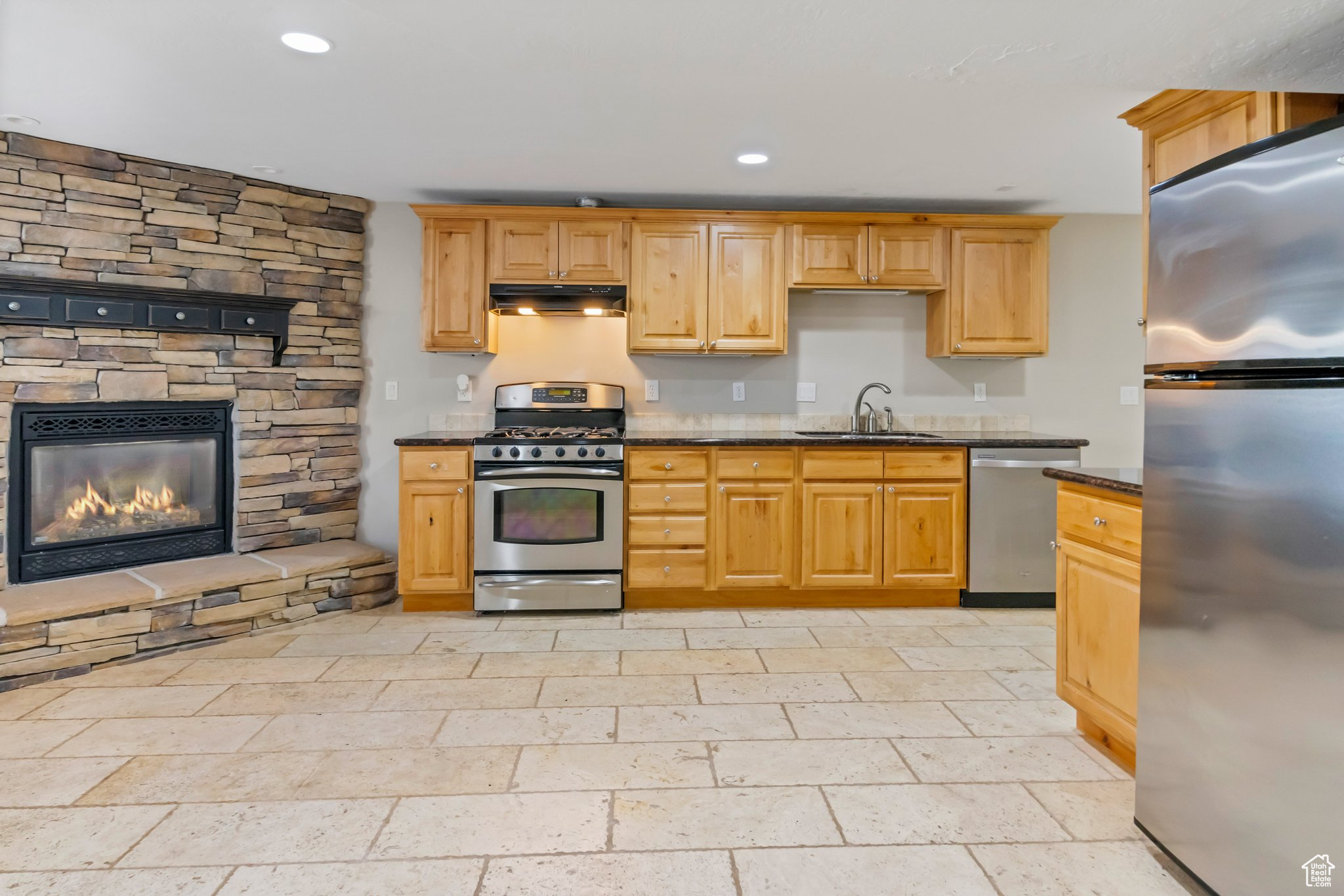 Kitchen with a stone fireplace, stainless steel appliances, light tile flooring, dark stone countertops, and sink