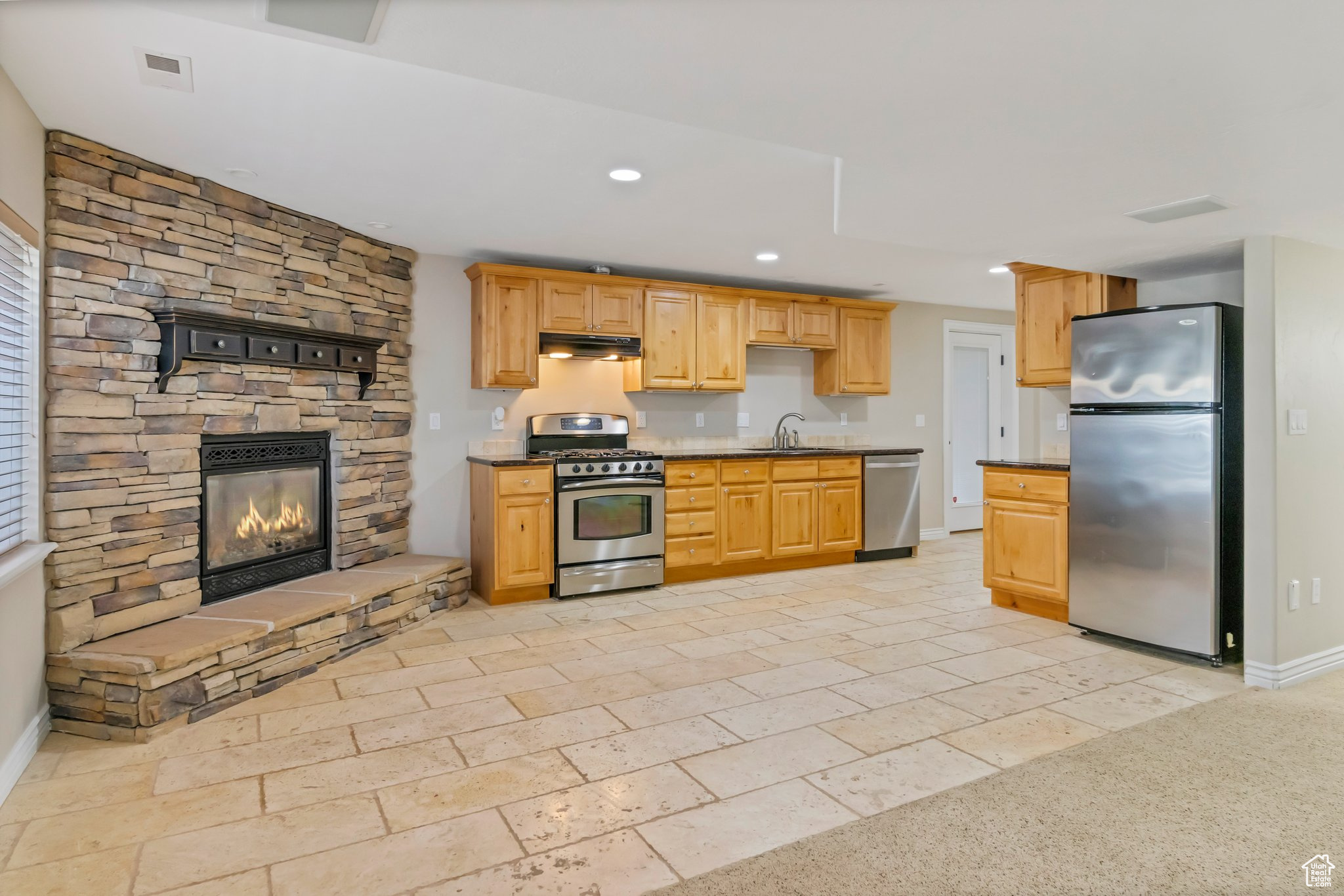 Kitchen with light brown cabinetry, sink, stainless steel appliances, and a stone fireplace