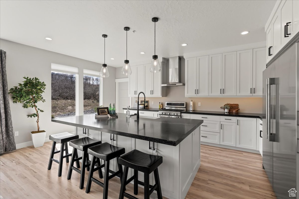 Kitchen with stainless steel appliances, wall chimney range hood, an island with sink, and decorative light fixtures