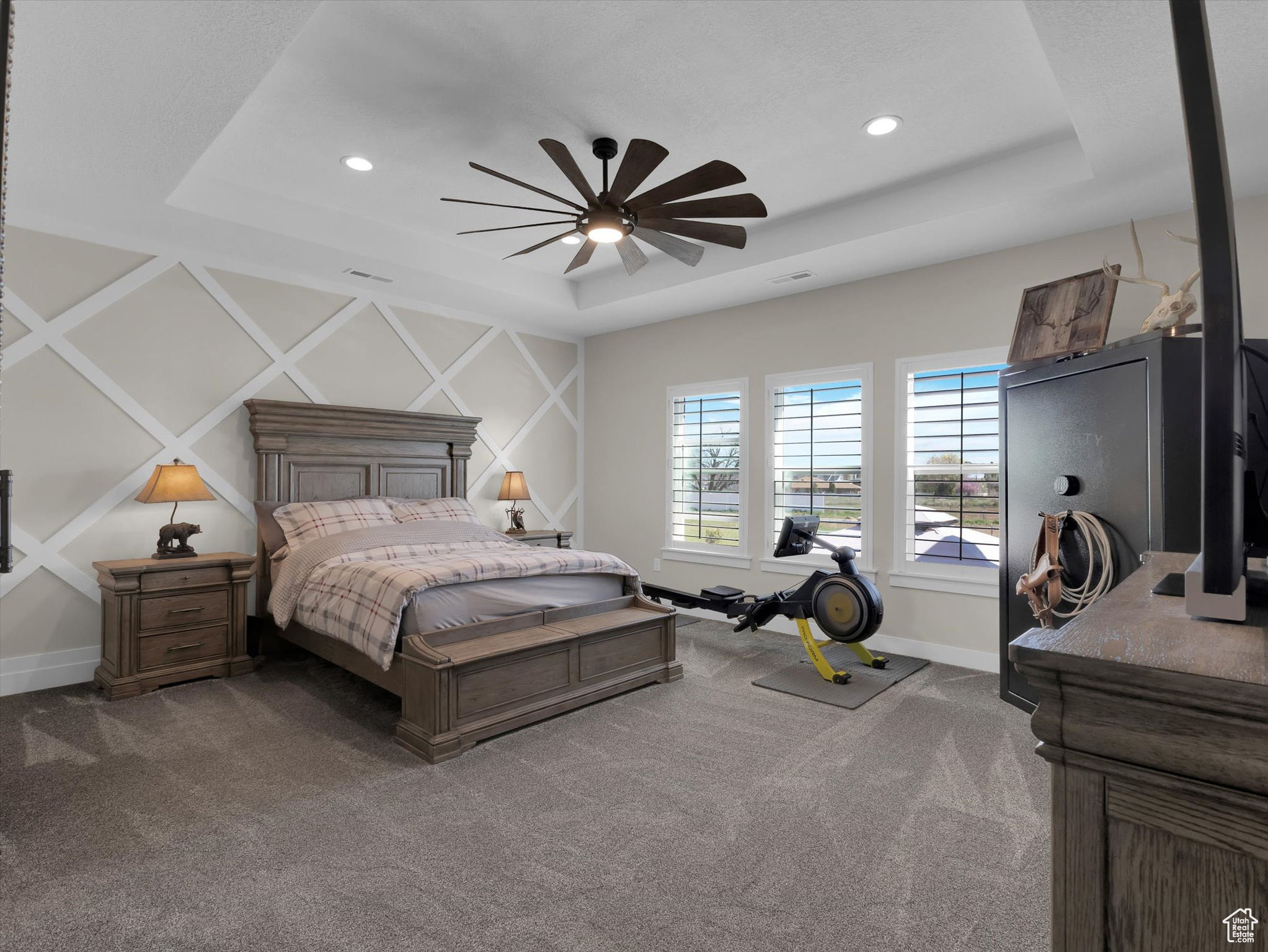 Primary bedroom featuring dark colored carpet, ceiling fan, wall feature and a tray ceiling