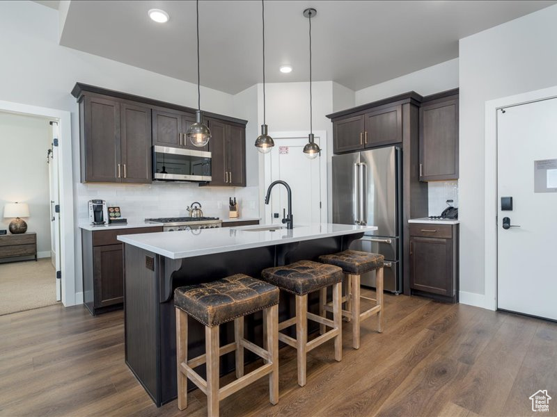 Kitchen with tasteful backsplash, dark wood-type flooring, decorative light fixtures, stainless steel appliances, and a kitchen island with sink