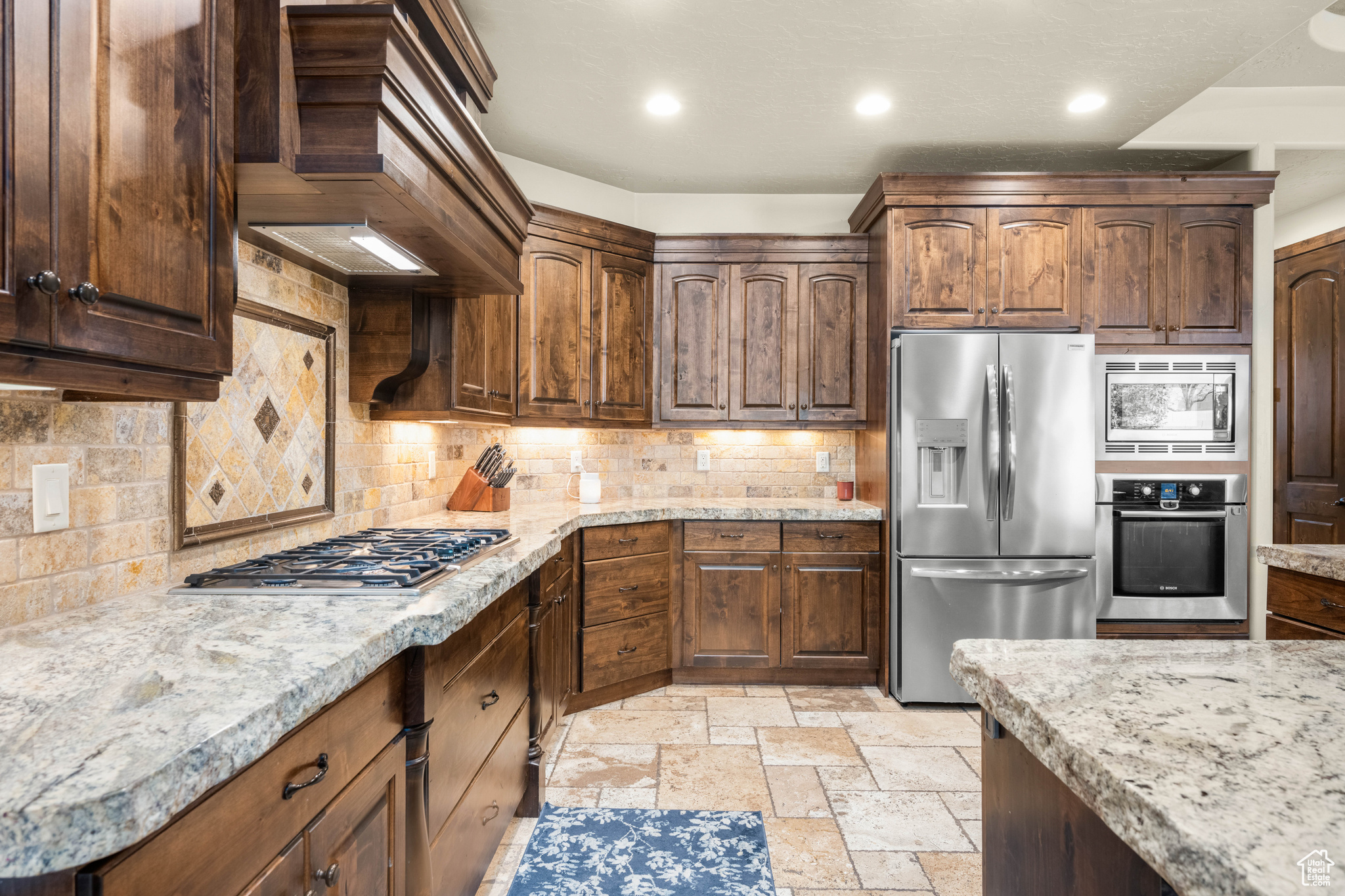 Kitchen with light stone counters, light tile floors, tasteful backsplash, stainless steel appliances, and custom range hood
