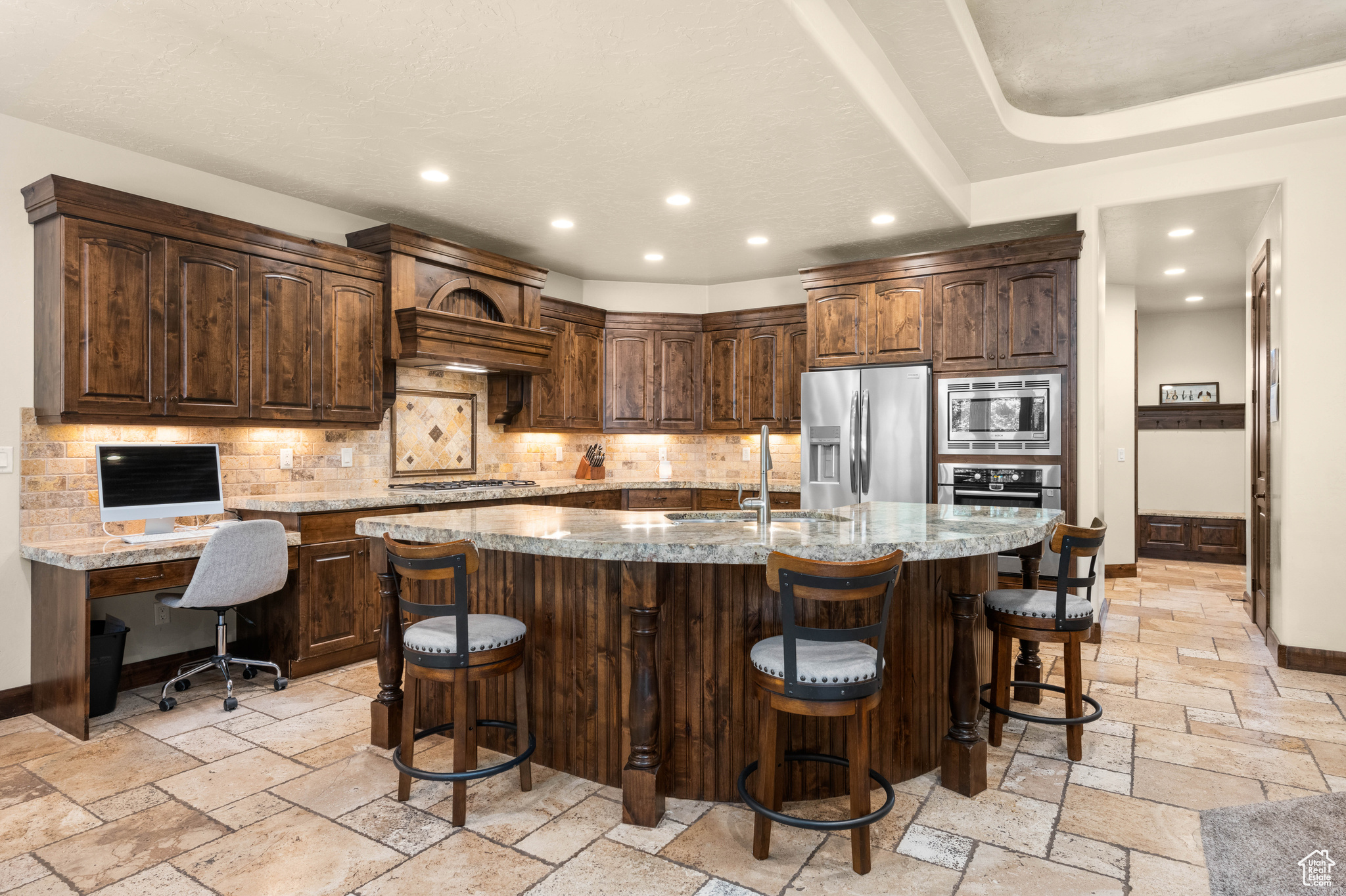Kitchen featuring light tile flooring, backsplash, appliances with stainless steel finishes, a center island with sink, and light stone counters