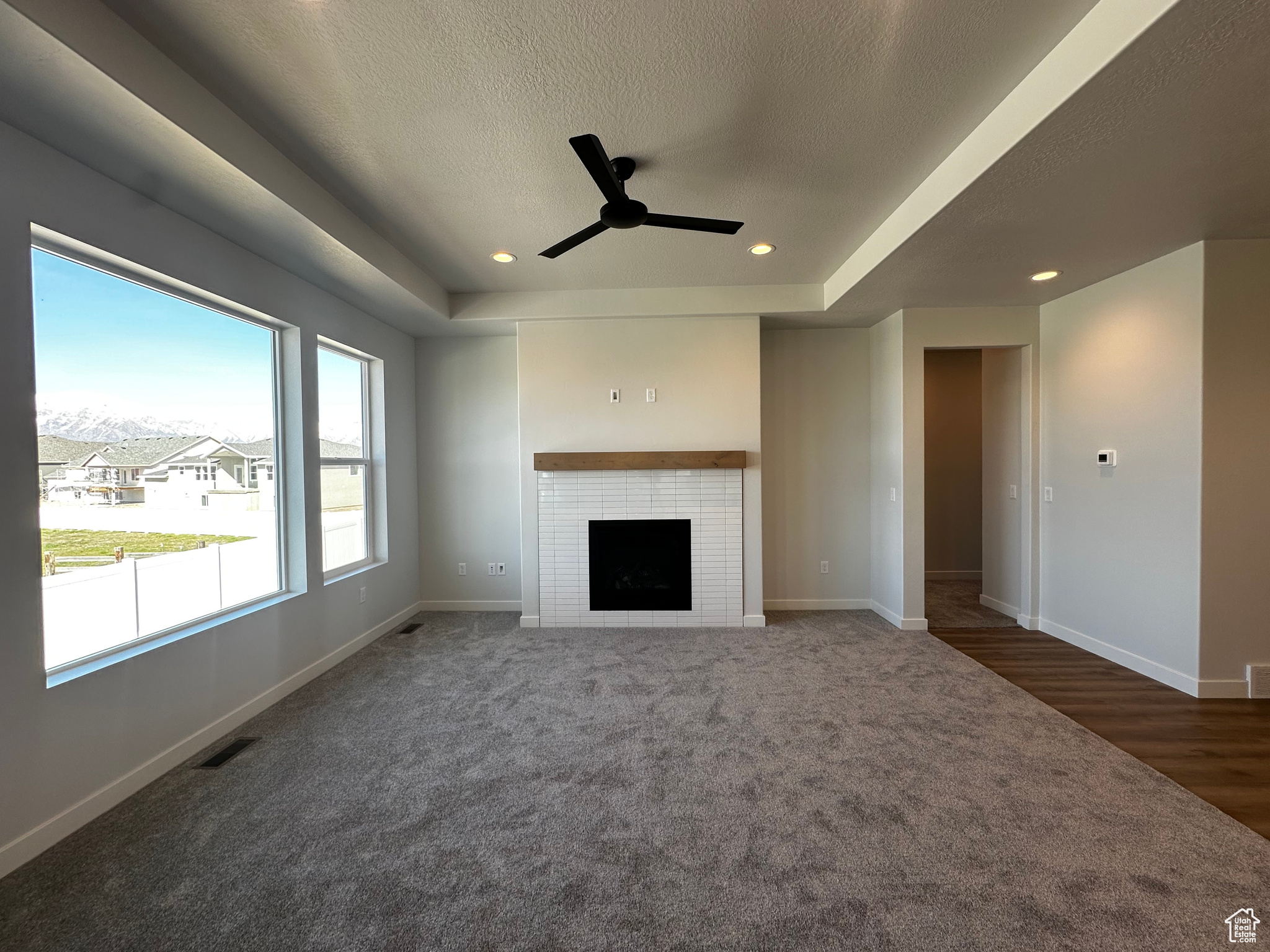 Unfurnished living room featuring a brick fireplace, dark colored carpet, a raised ceiling, ceiling fan, and a textured ceiling