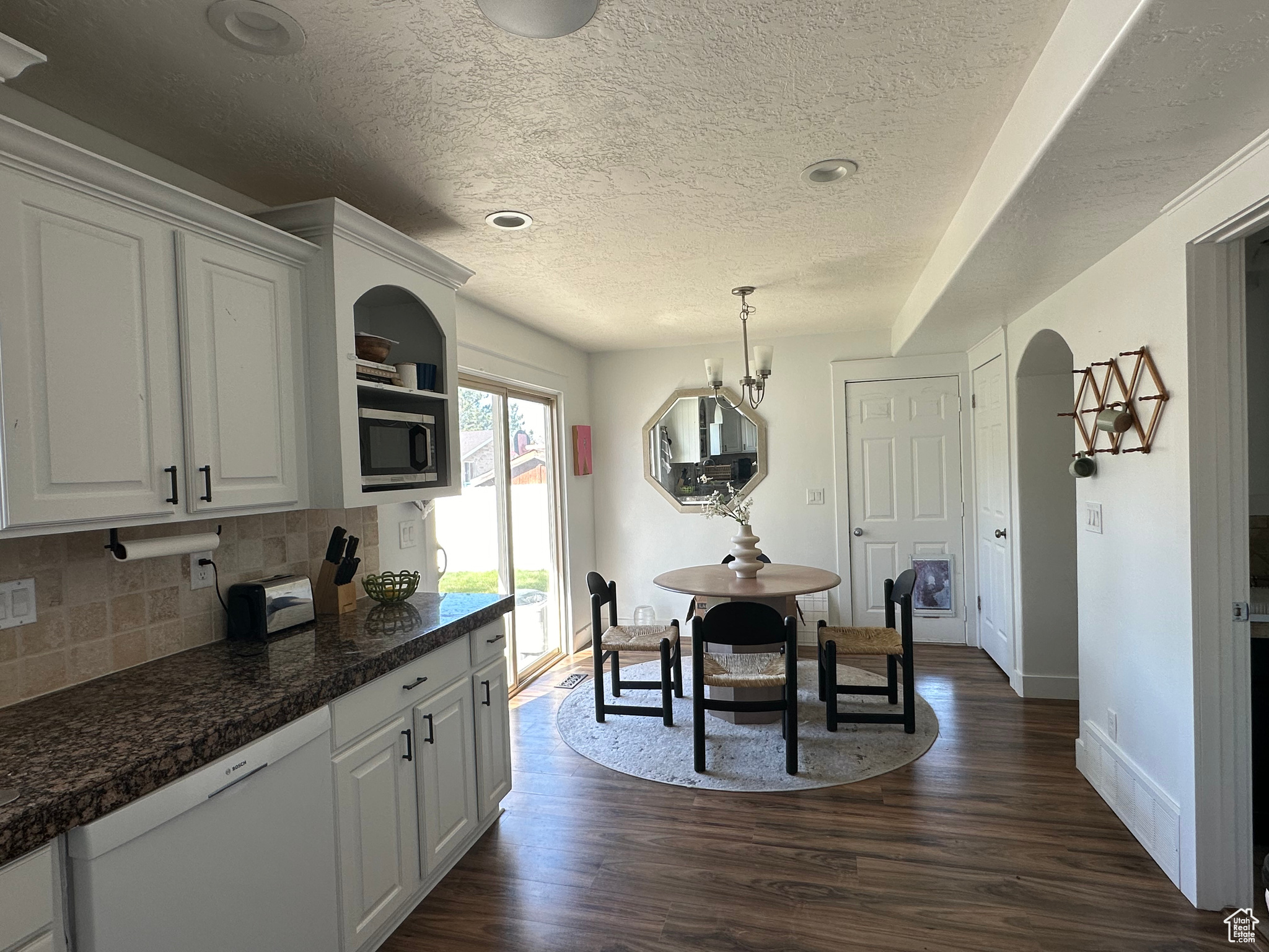 Kitchen with hanging light fixtures, dark wood-type flooring, white cabinetry, backsplash, and dishwasher