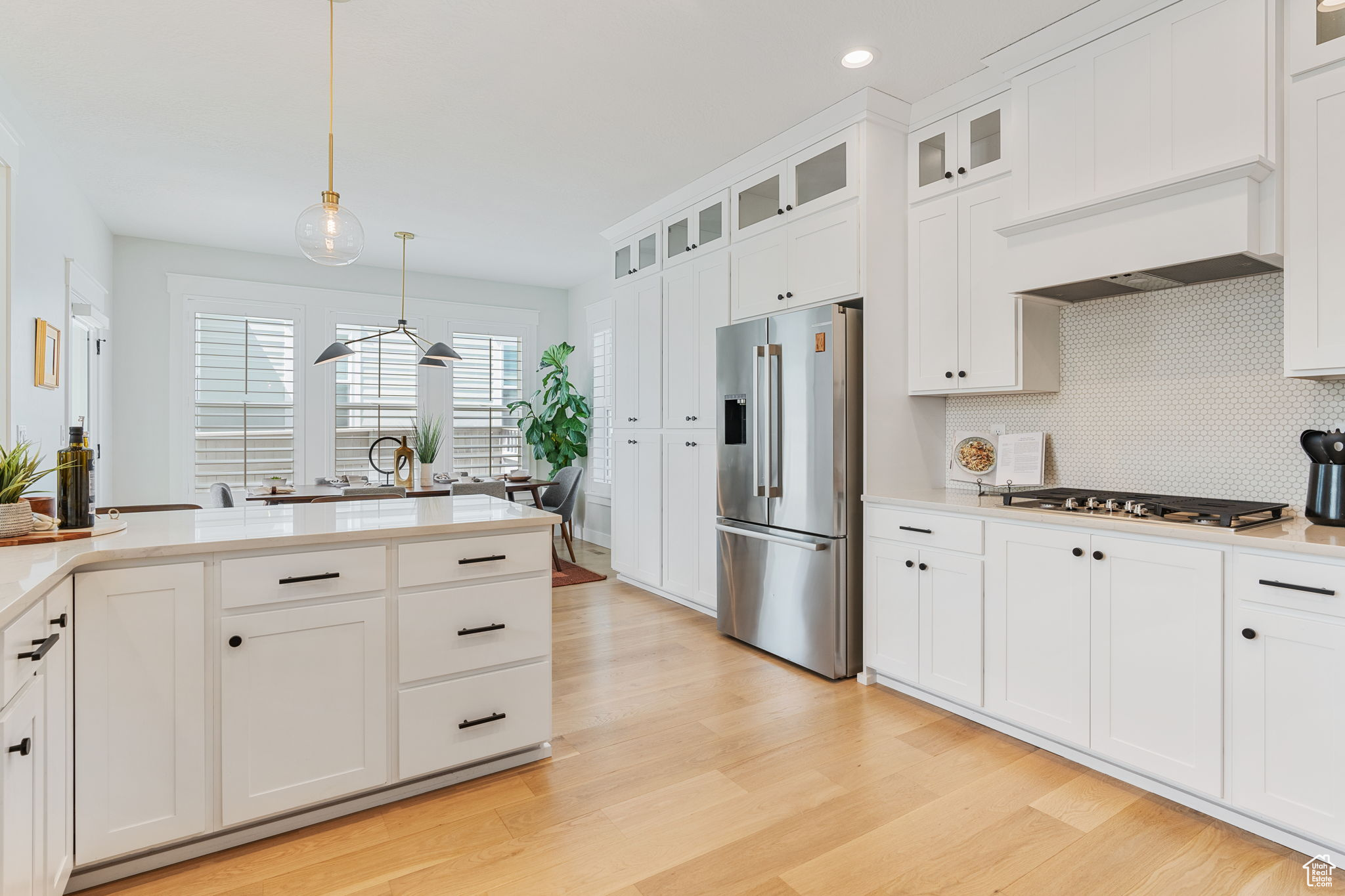 Kitchen featuring contemporary light fixtures, light oak hardwood, white cabinetry, backsplash, double oven, gas cooktop, vent hood, and stainless steel appliances.