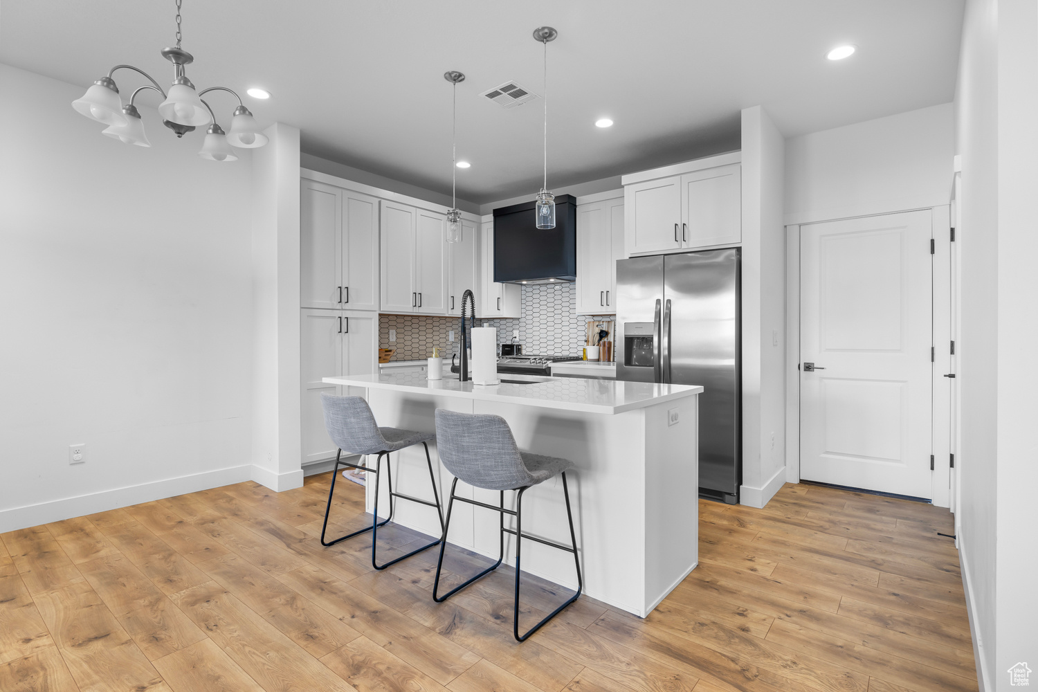 Kitchen with backsplash, light hardwood / wood-style flooring, stainless steel fridge, a kitchen island with sink, and pendant lighting
