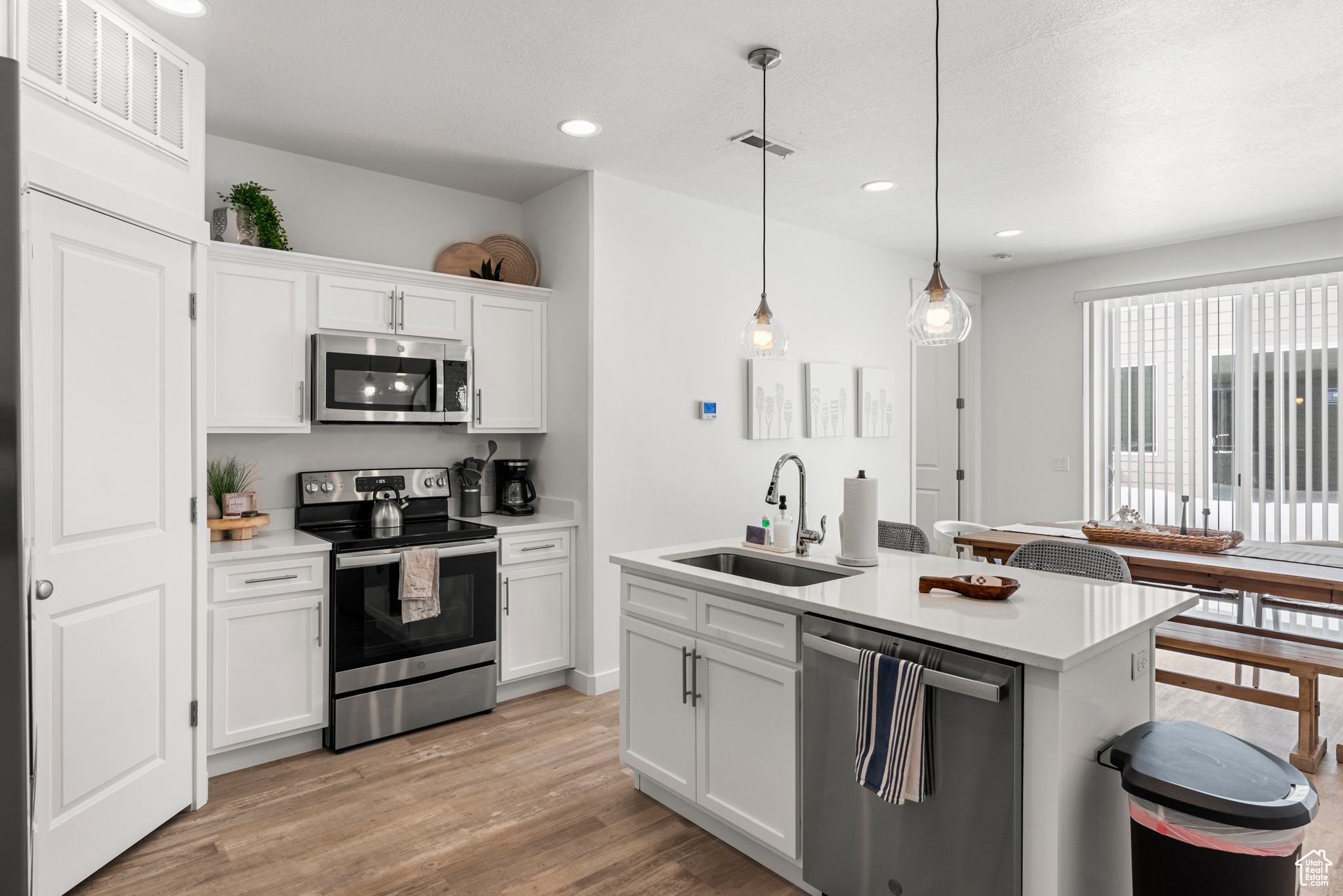 Kitchen with appliances with stainless steel finishes, light hardwood / wood-style flooring, white cabinetry, hanging light fixtures, and an island with sink