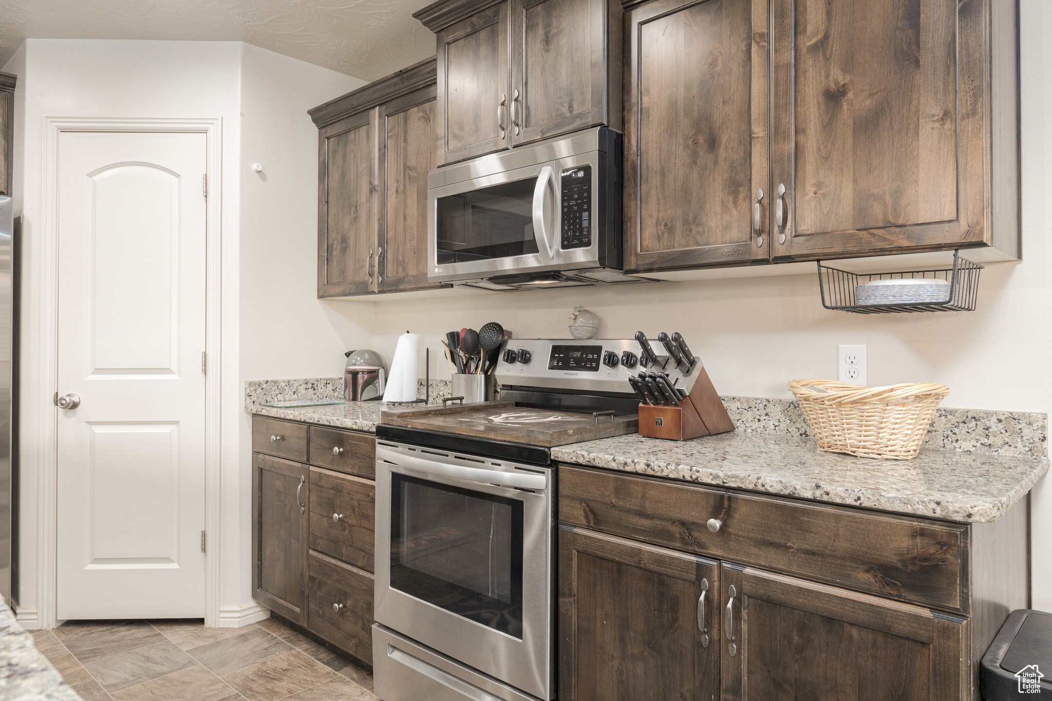 Kitchen featuring stainless steel appliances, dark brown cabinetry, light tile floors, and light stone countertops