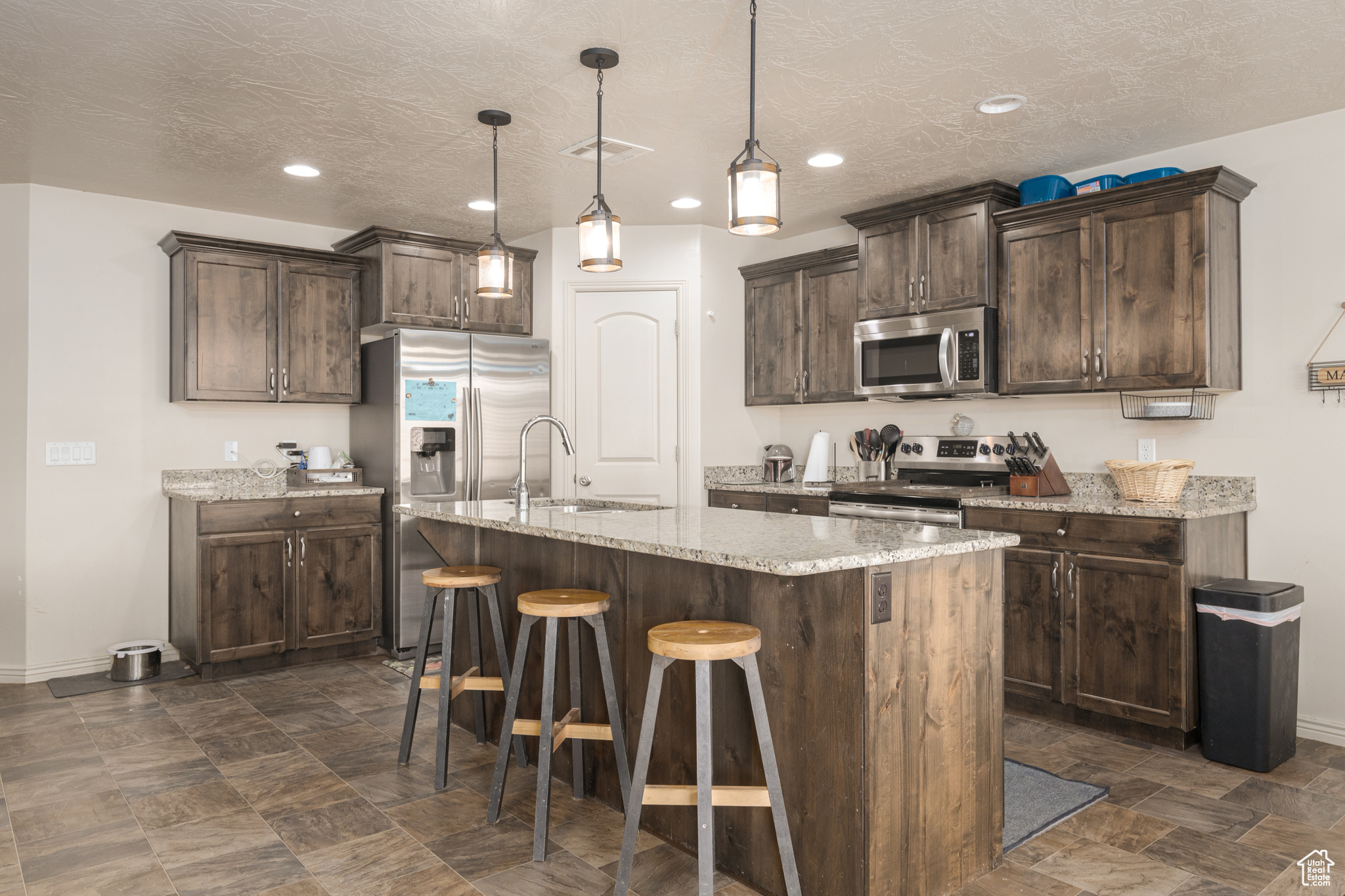 Kitchen featuring dark brown cabinetry, appliances with stainless steel finishes, sink, decorative light fixtures, and a kitchen island with sink