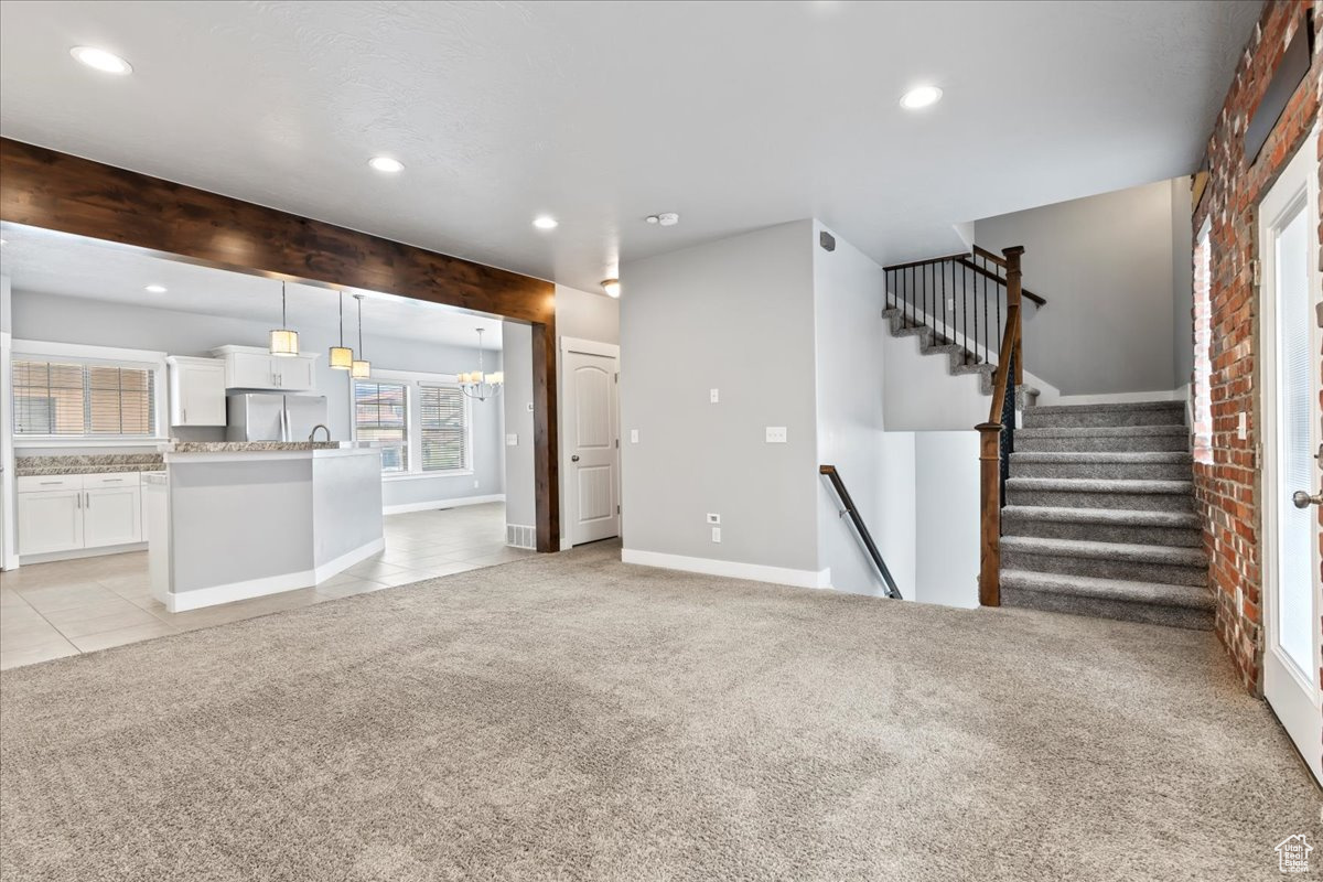 Unfurnished living room featuring light carpet, sink, a notable chandelier, beam ceiling, and brick wall