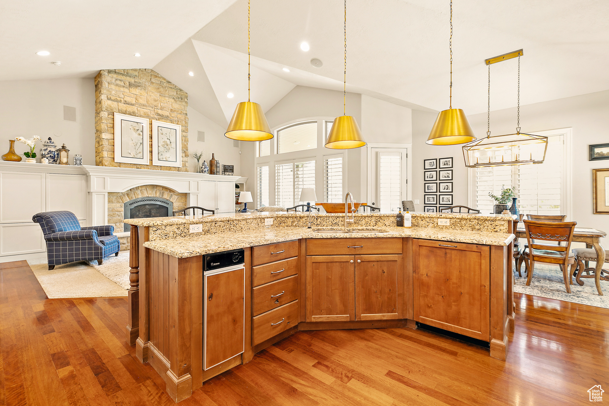 Kitchen featuring a stone fireplace, hanging light fixtures, a kitchen island with sink, light stone counters, and hardwood / wood-style floors