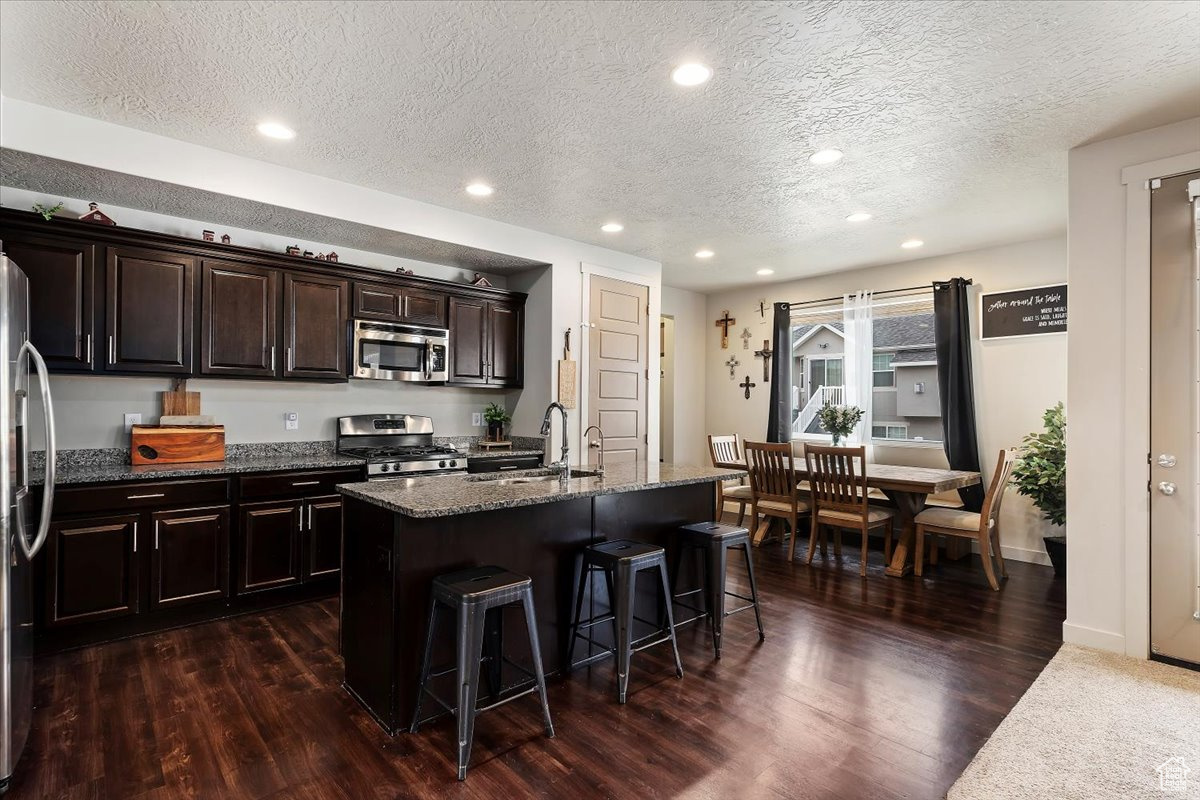 Kitchen with stainless steel appliances, a center island with sink, dark stone counters, sink, and dark hardwood / wood-style flooring
