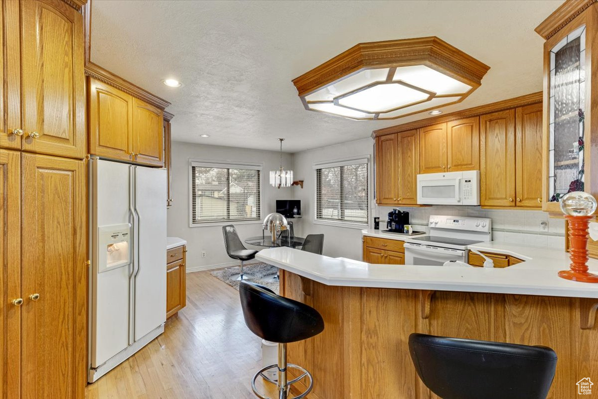 Kitchen with light hardwood / wood-style floors, white appliances, decorative light fixtures, kitchen peninsula, and a kitchen breakfast bar