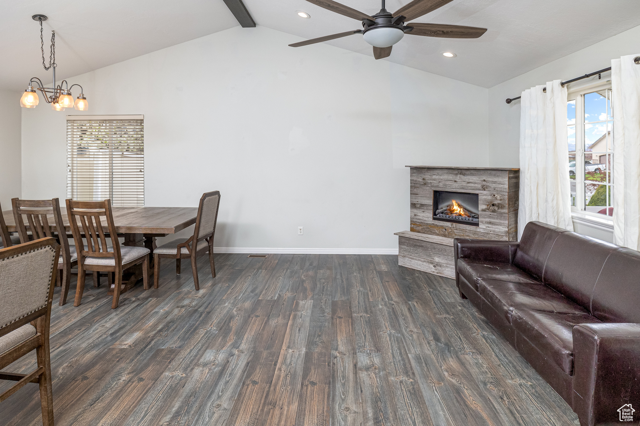 Living room featuring ceiling fan with notable chandelier, dark wood-type flooring, vaulted ceiling with beams, and a fireplace