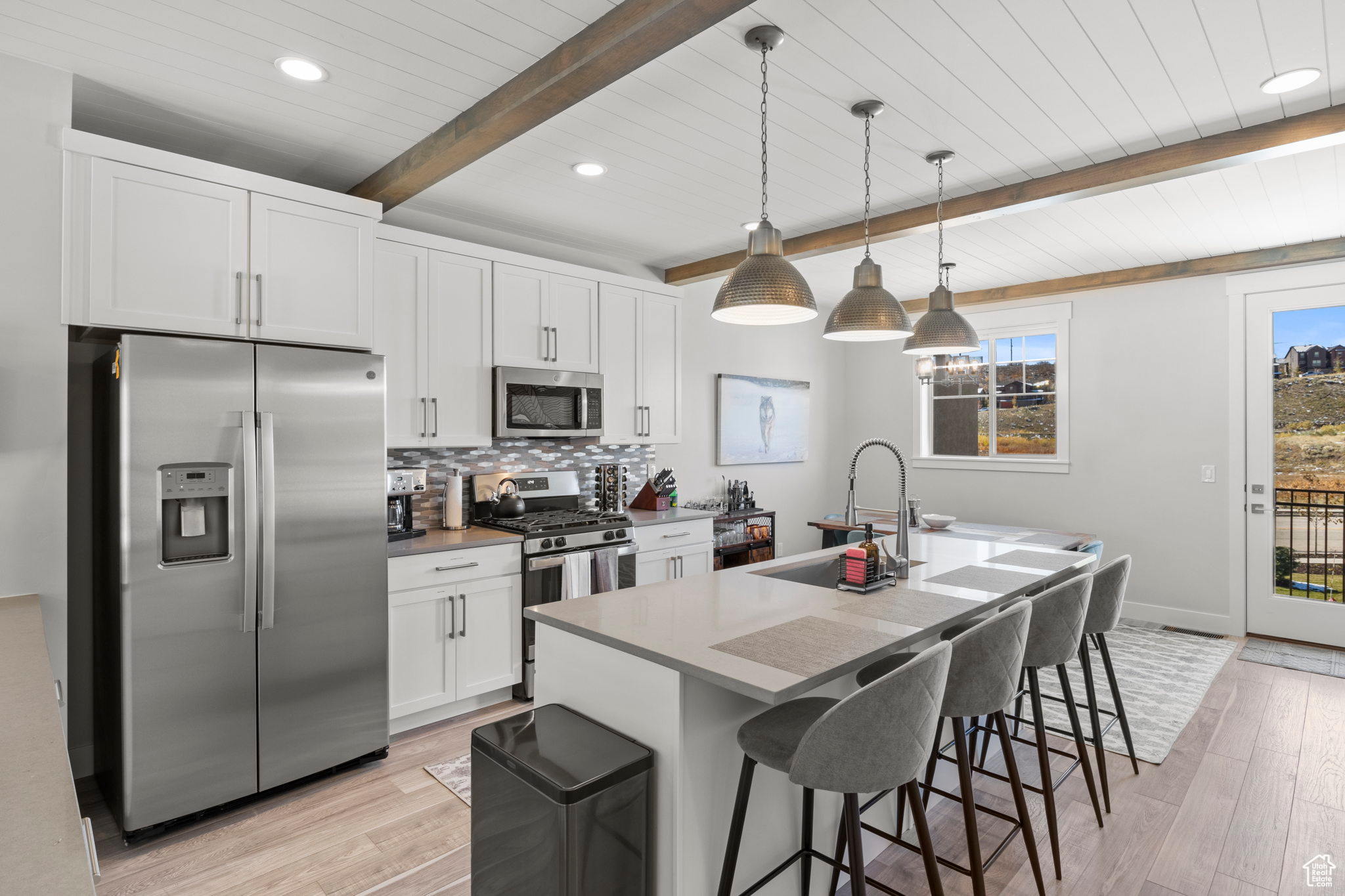 Kitchen with appliances with stainless steel finishes, light wood-type flooring, hanging light fixtures, beam ceiling, and an island with sink