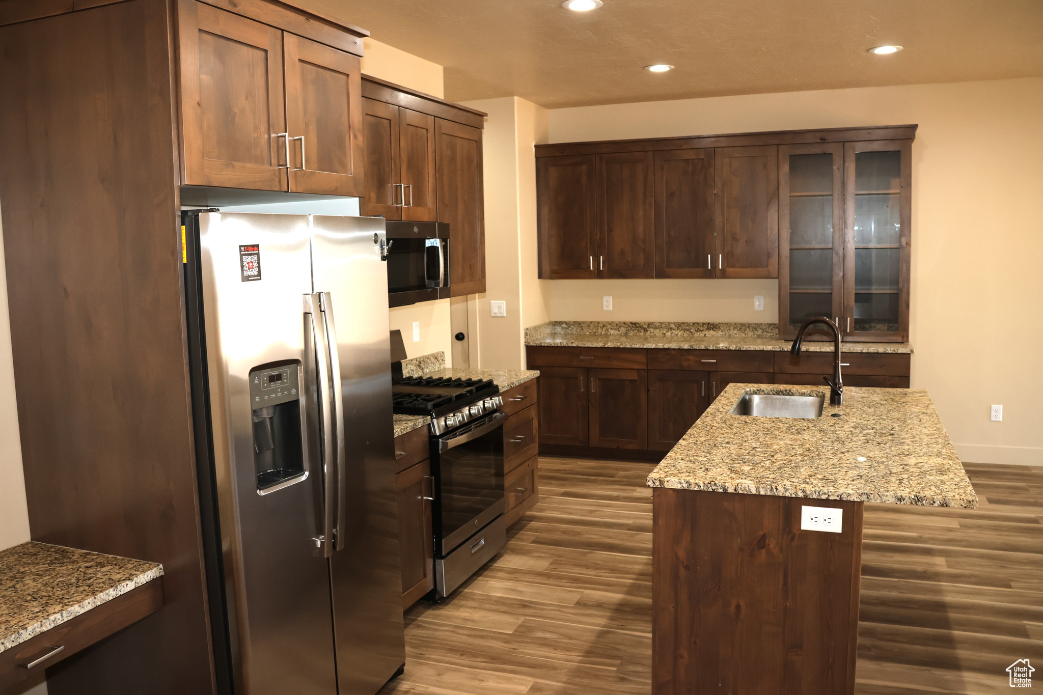 Kitchen with wood-type flooring, light stone countertops, appliances with stainless steel finishes, sink, and an island with sink