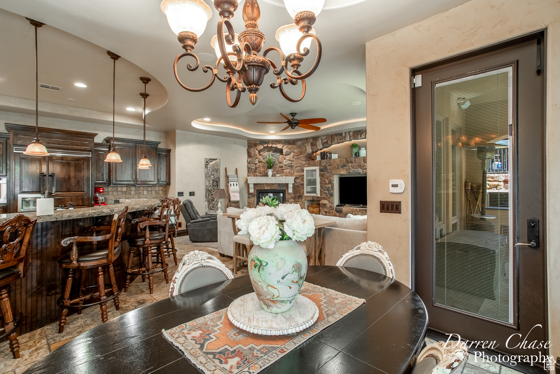 Tiled dining space featuring ceiling fan with notable chandelier and a stone fireplace