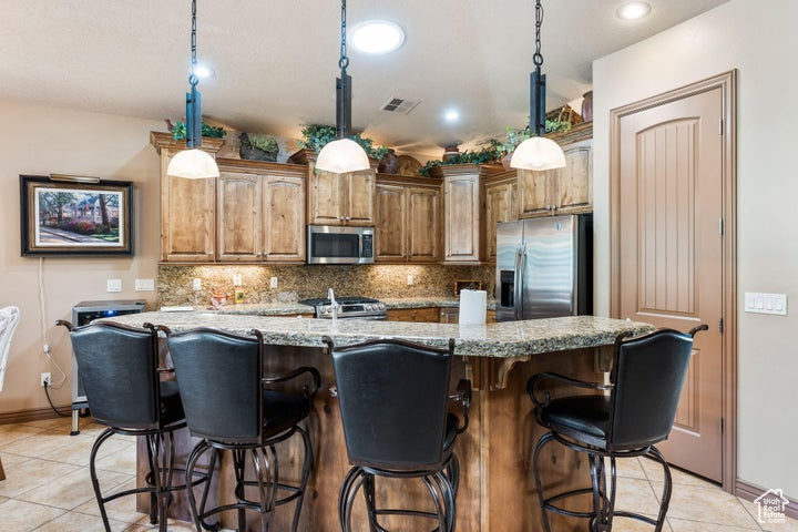 Kitchen featuring hanging light fixtures, light tile flooring, backsplash, stainless steel appliances, and an island with sink