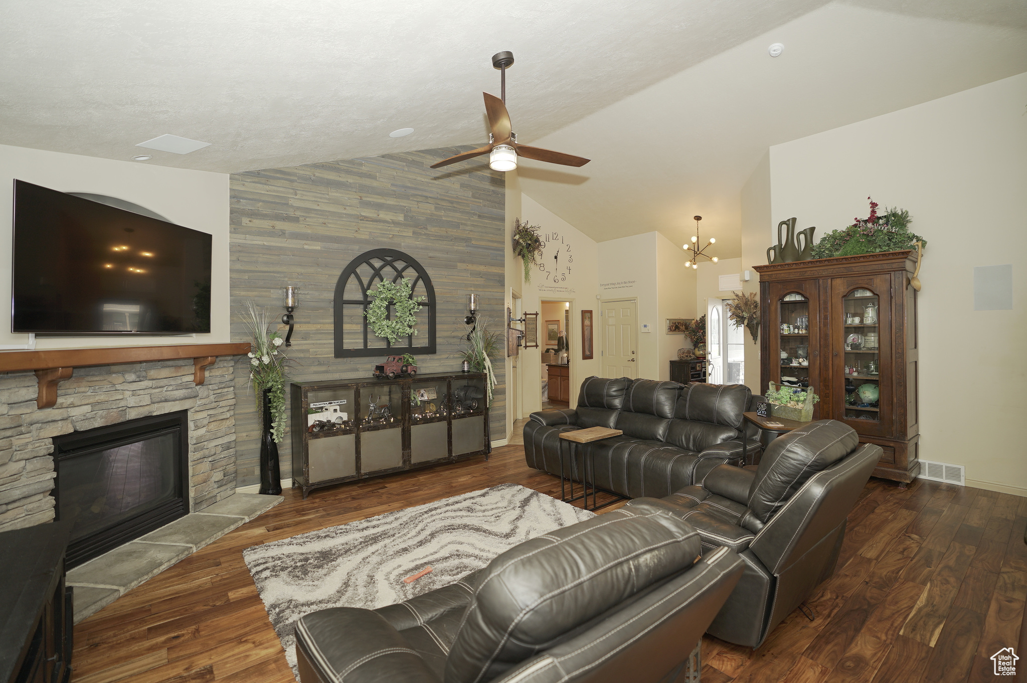 Living room with a stone fireplace, high vaulted ceiling, ceiling fan with notable chandelier, dark wood-type flooring, and wood walls