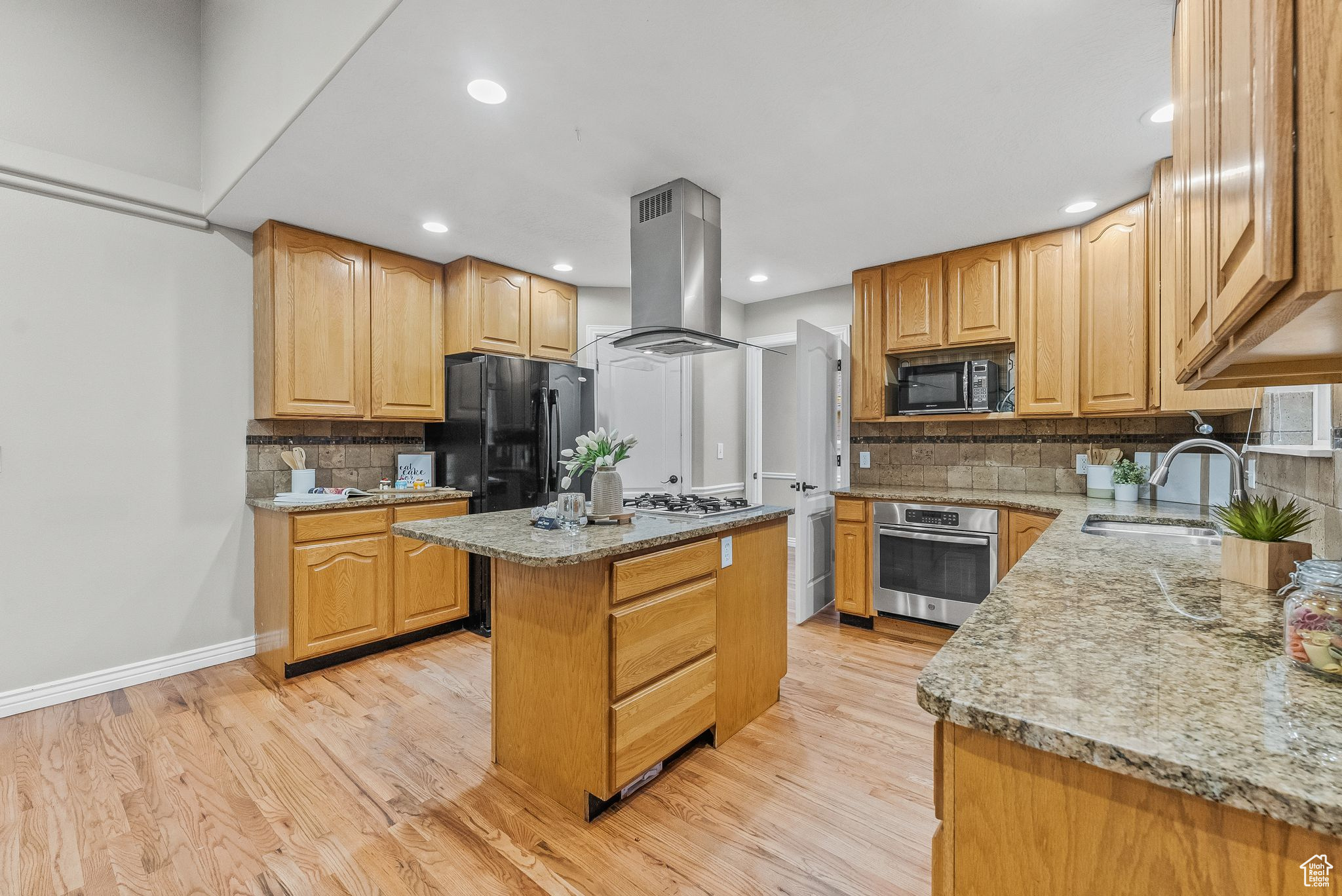Kitchen with island range hood, black appliances, tasteful backsplash, sink, and light wood-type flooring