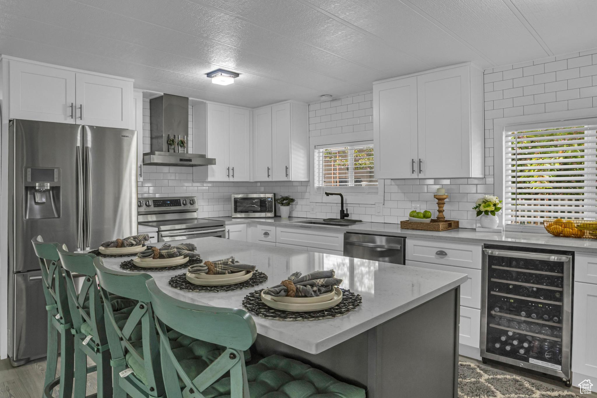 Kitchen with wall chimney exhaust hood, wine cooler, stainless steel appliances, and white cabinets