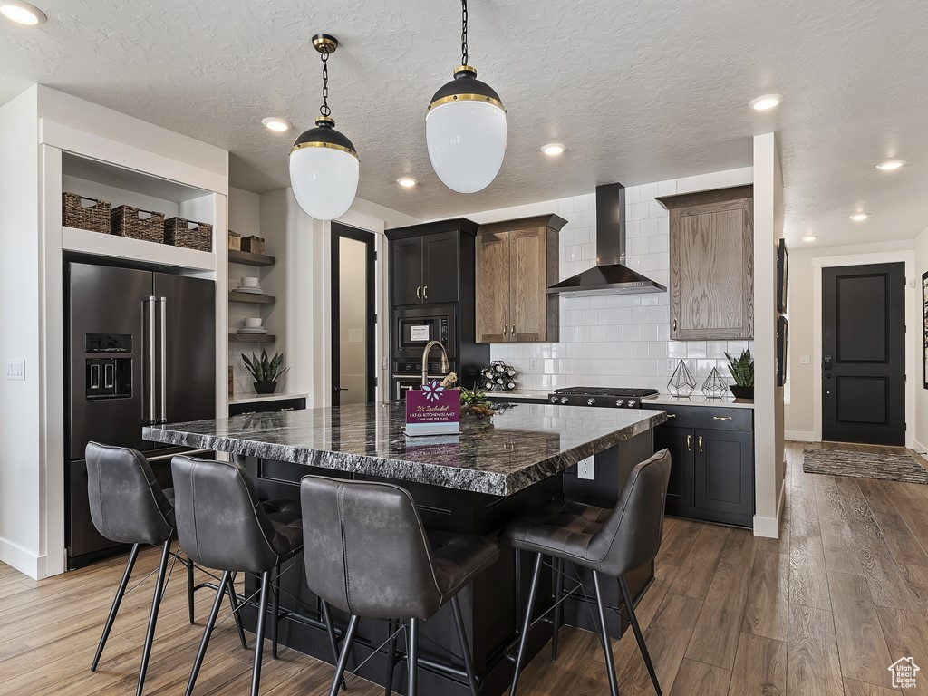 Kitchen featuring wood-type flooring, black appliances, backsplash, wall chimney range hood, and an island with sink