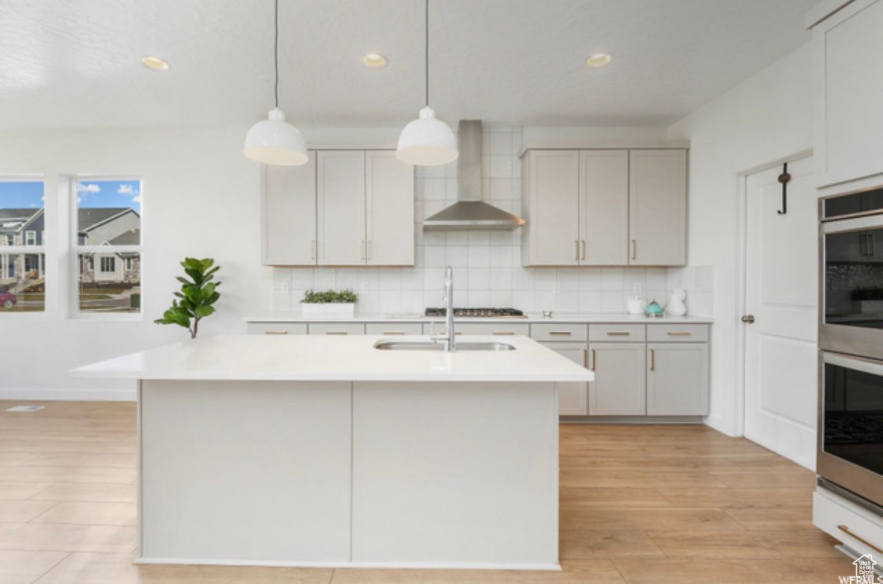 Kitchen featuring backsplash, light hardwood / wood-style floors, an island with sink, wall chimney exhaust hood, and pendant lighting