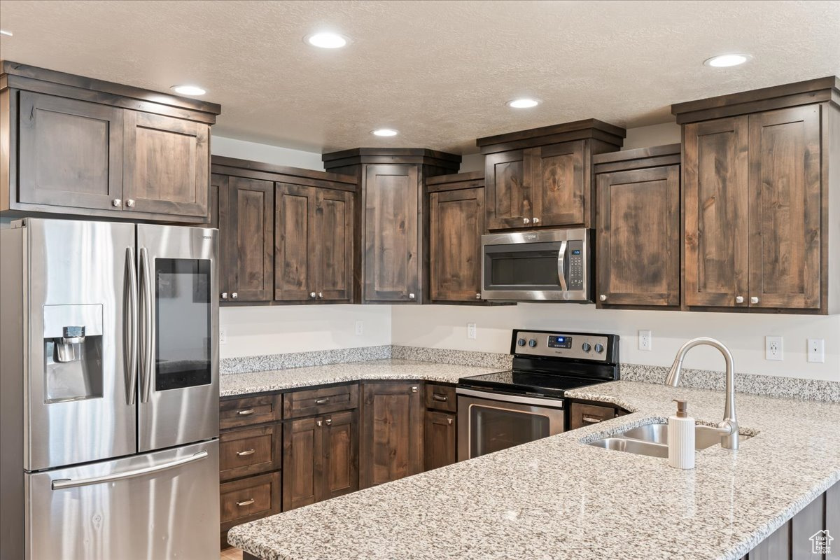 Kitchen with light stone countertops, kitchen peninsula, stainless steel appliances, sink, and dark brown cabinetry