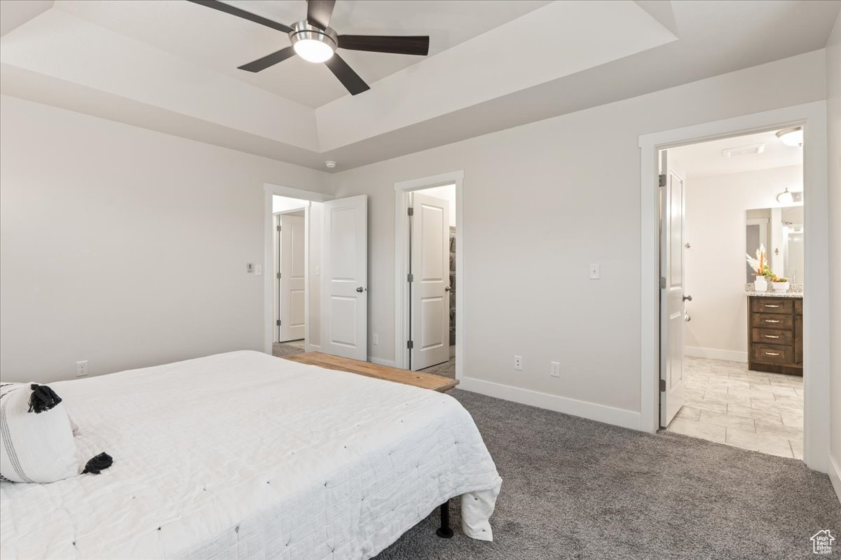 Primary bedroom featuring ensuite bath, ceiling fan, a tray ceiling, and light colored carpet