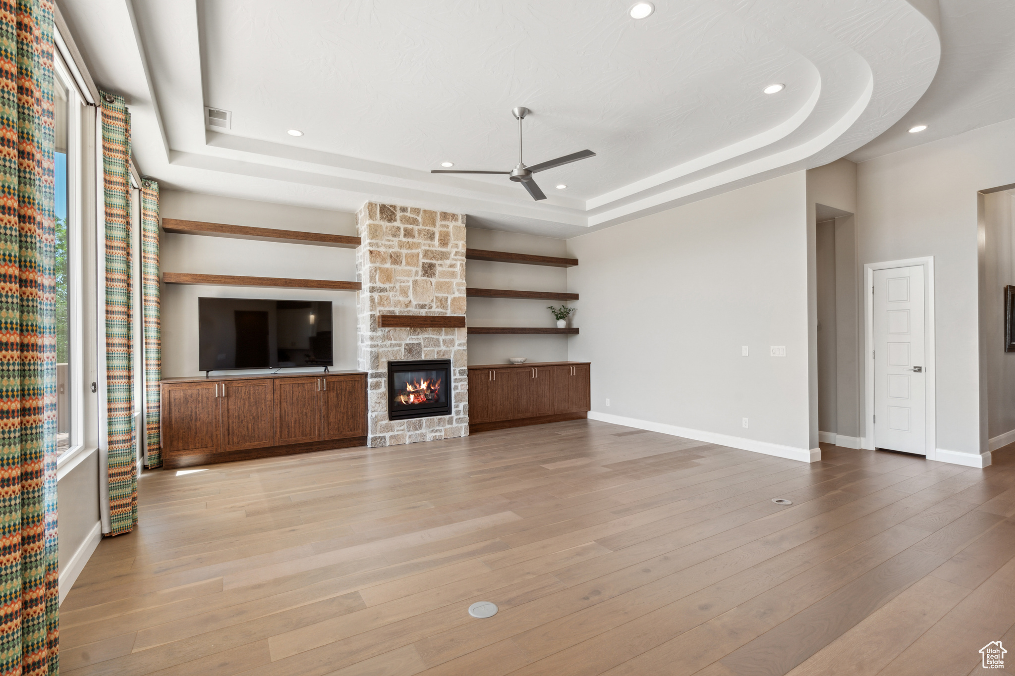Unfurnished living room featuring a raised ceiling, ceiling fan, light hardwood / wood-style floors, and a stone fireplace