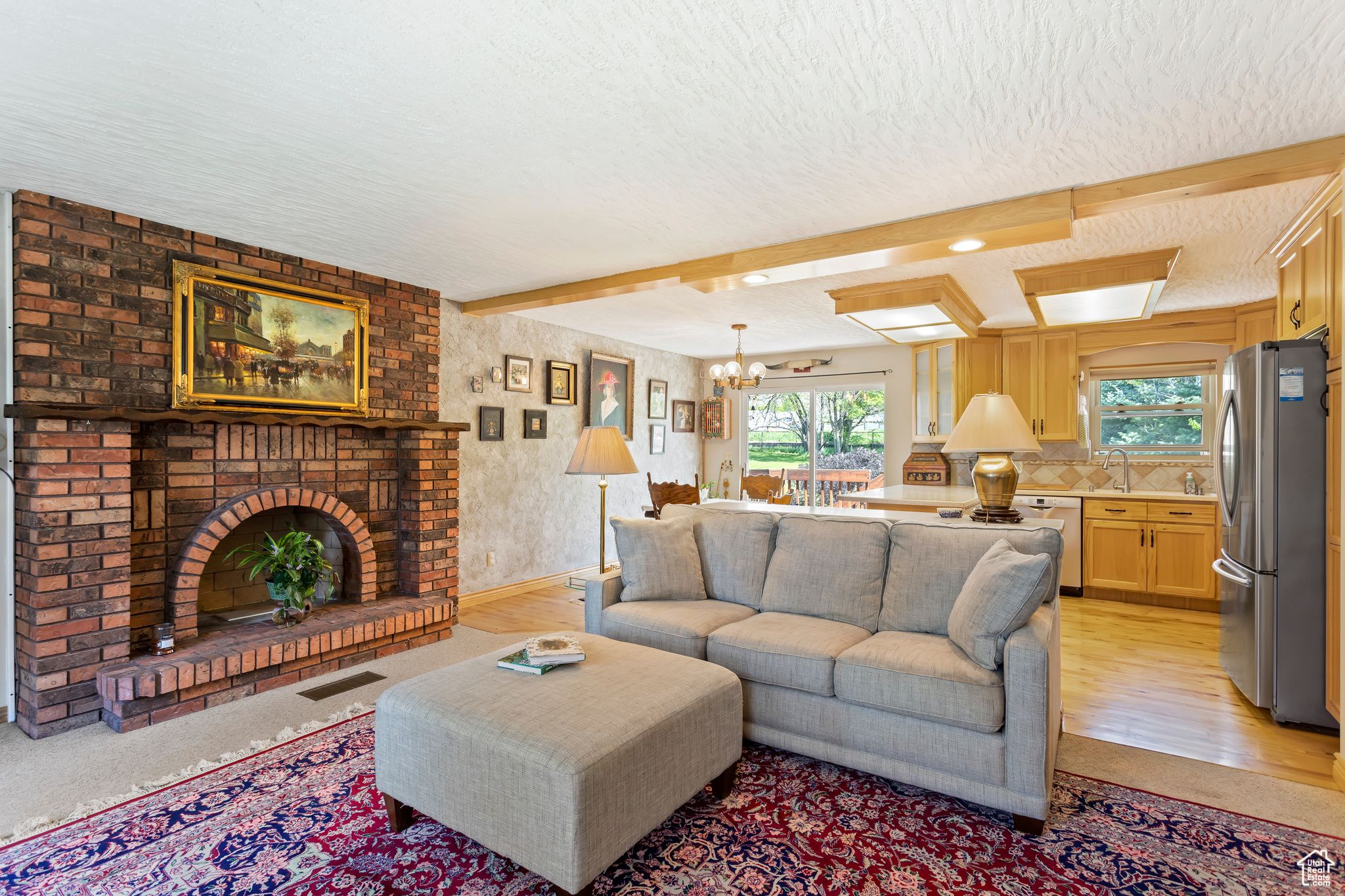 Carpeted living room with an inviting chandelier, beam ceiling, a healthy amount of sunlight, and a brick fireplace