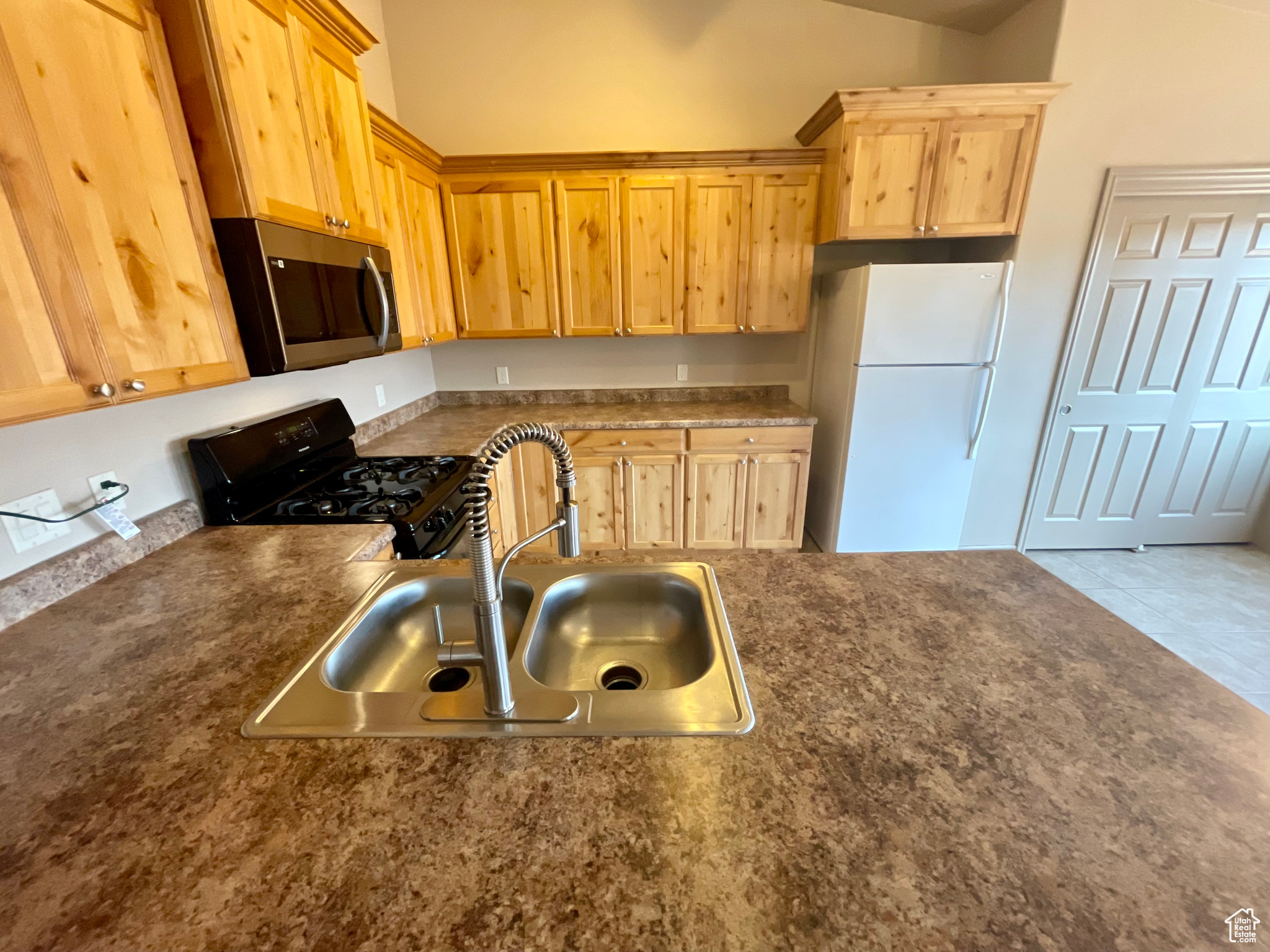 Kitchen with light brown cabinetry, black gas range oven, light tile patterned floors, white fridge, and sink