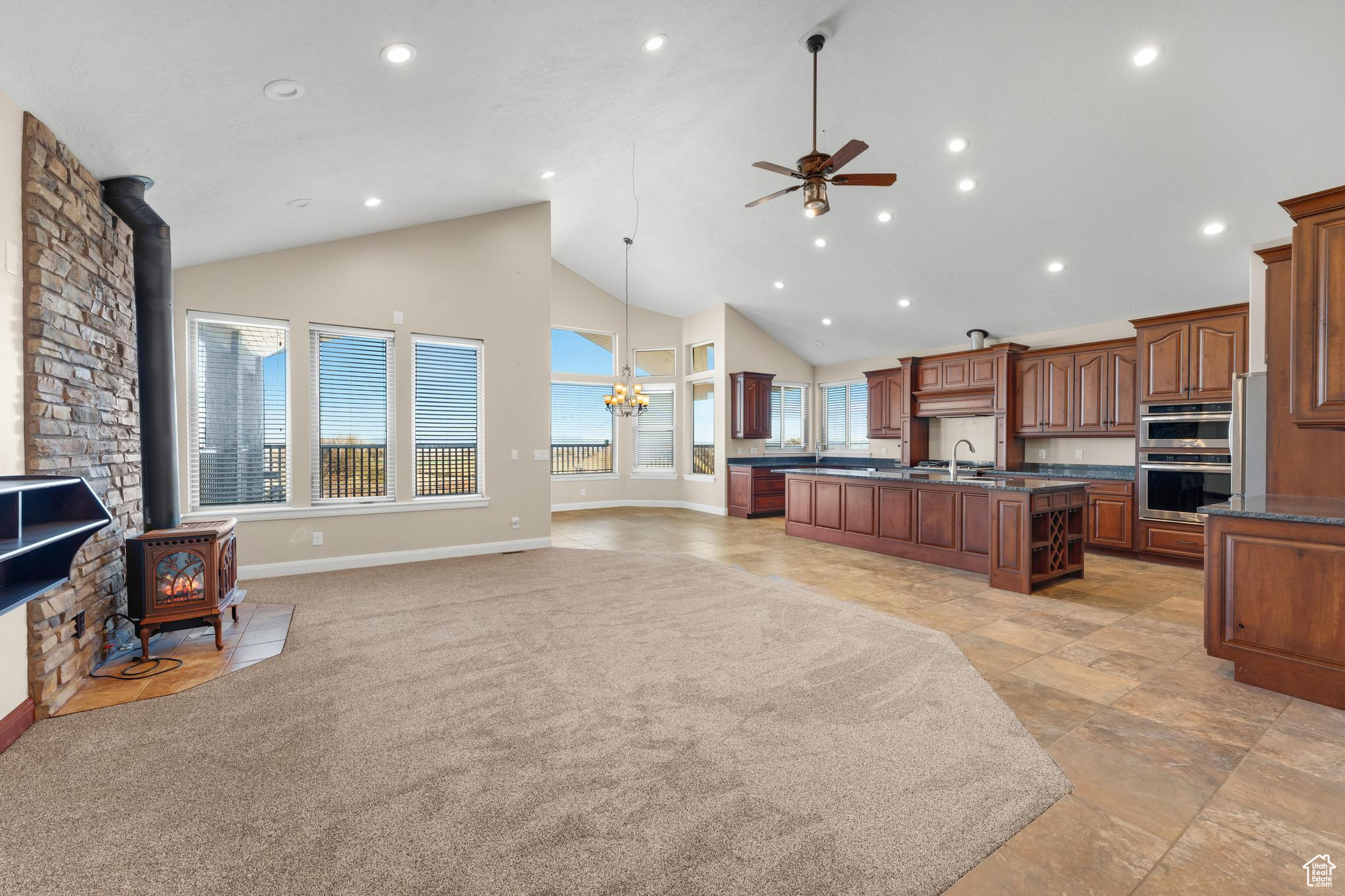 Kitchen with ceiling fan with notable chandelier, stainless steel double oven, light tile floors, a wood stove, and a center island with sink