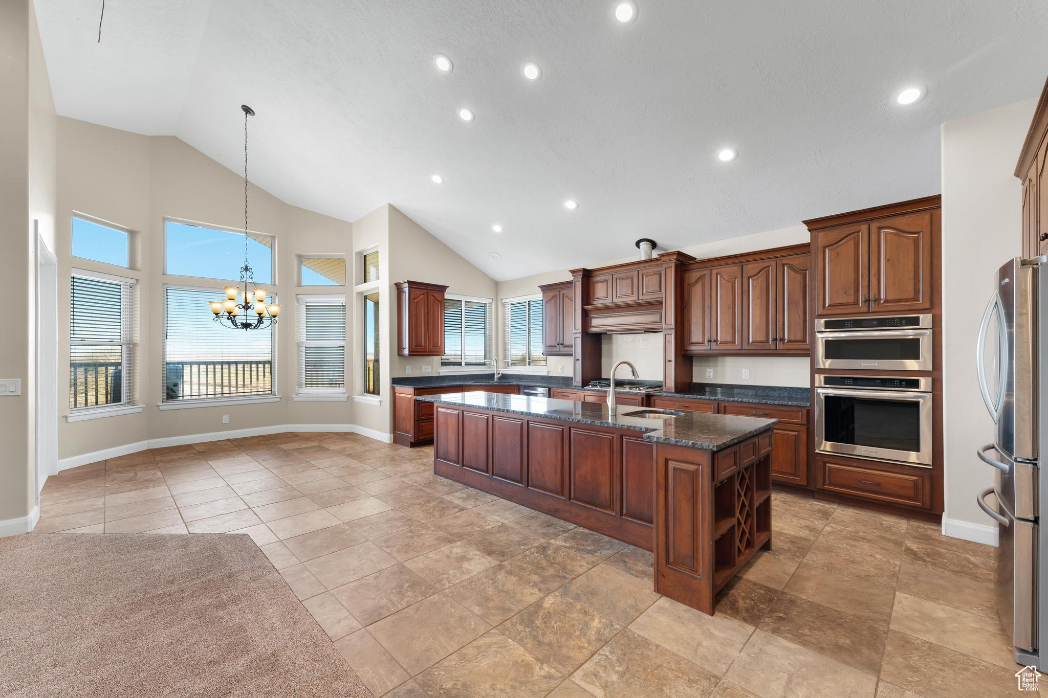 Kitchen with light tile floors, dark stone counters, a center island with sink, stainless steel appliances, and an inviting chandelier