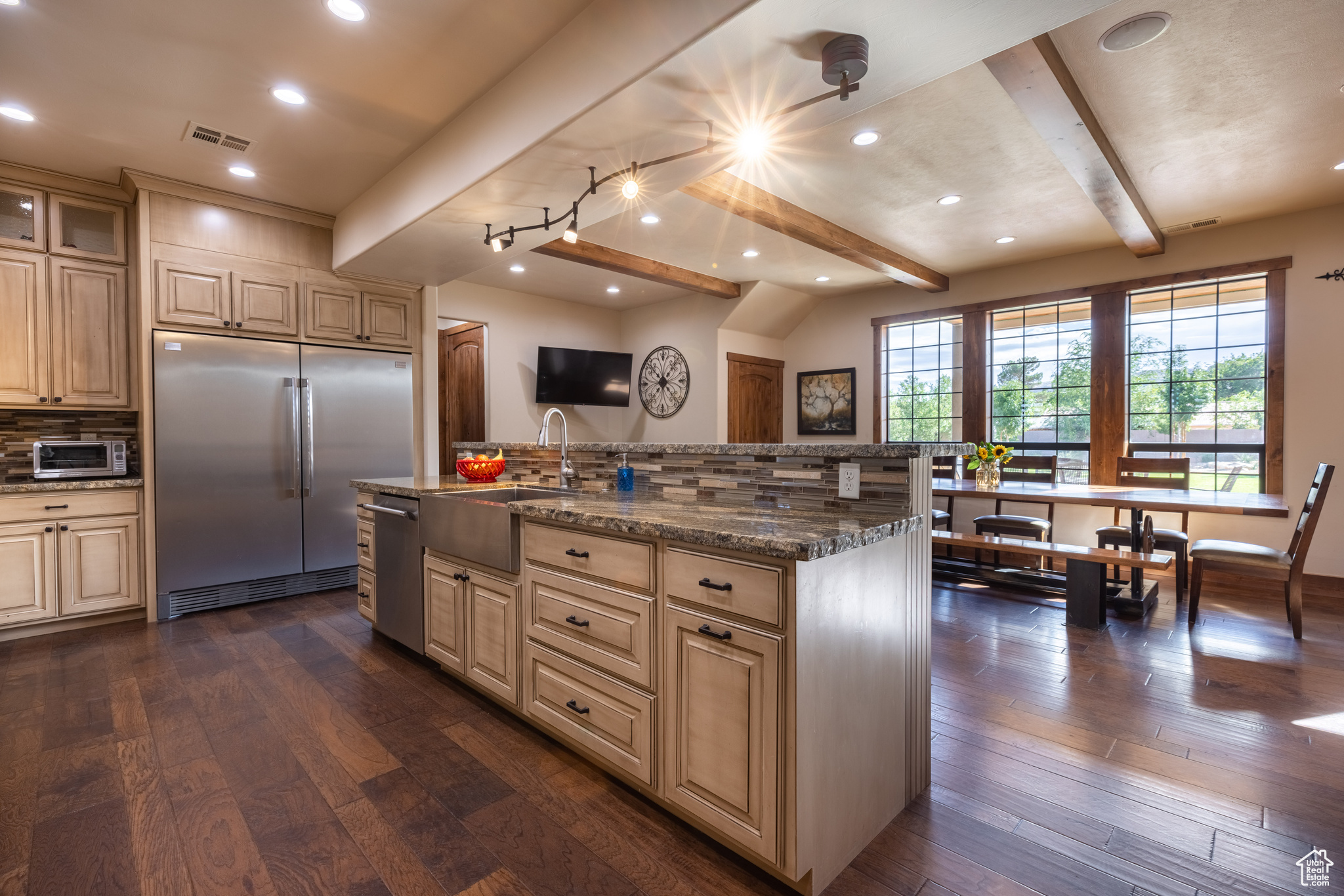 Kitchen with backsplash, stainless steel appliances, beam ceiling, dark wood-type flooring, and an island with sink