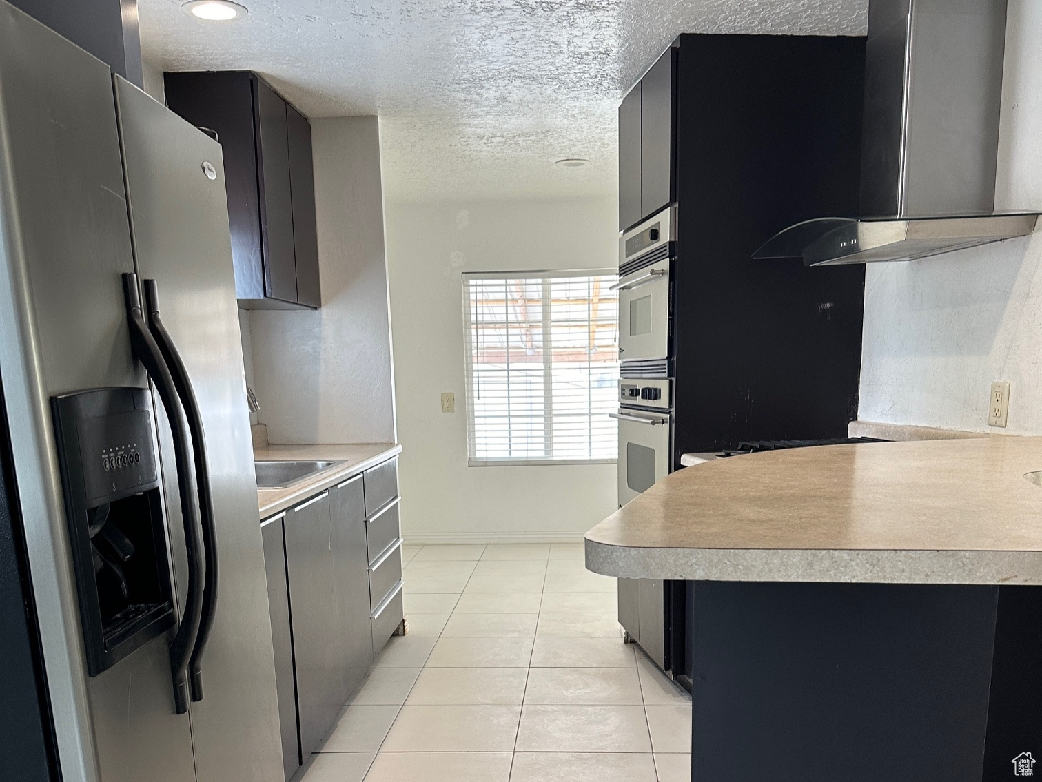 Kitchen featuring wall chimney range hood, a textured ceiling, appliances with stainless steel finishes, sink, and light tile floors