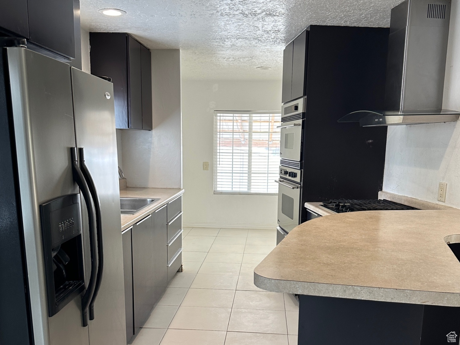 Kitchen with a textured ceiling, light tile floors, wall chimney exhaust hood, sink, and appliances with stainless steel finishes