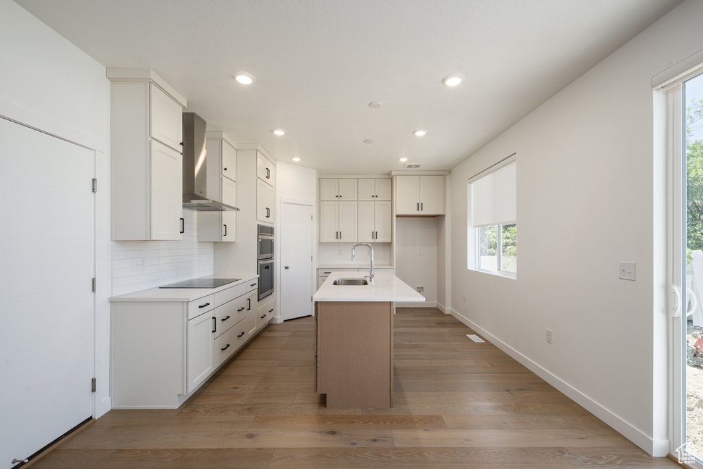 Kitchen with light hardwood / wood-style flooring, plenty of natural light, wall chimney exhaust hood, and a center island with sink