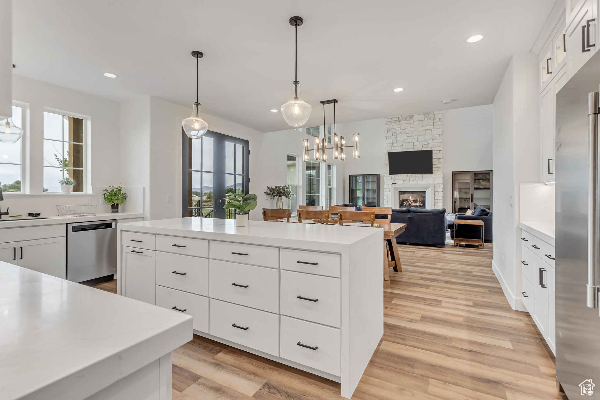 Kitchen with light hardwood / wood-style flooring, white cabinetry with roll trays in the lower cabinets, a custom surround stone fireplace, appliances with stainless steel finishes, and pendant lighting