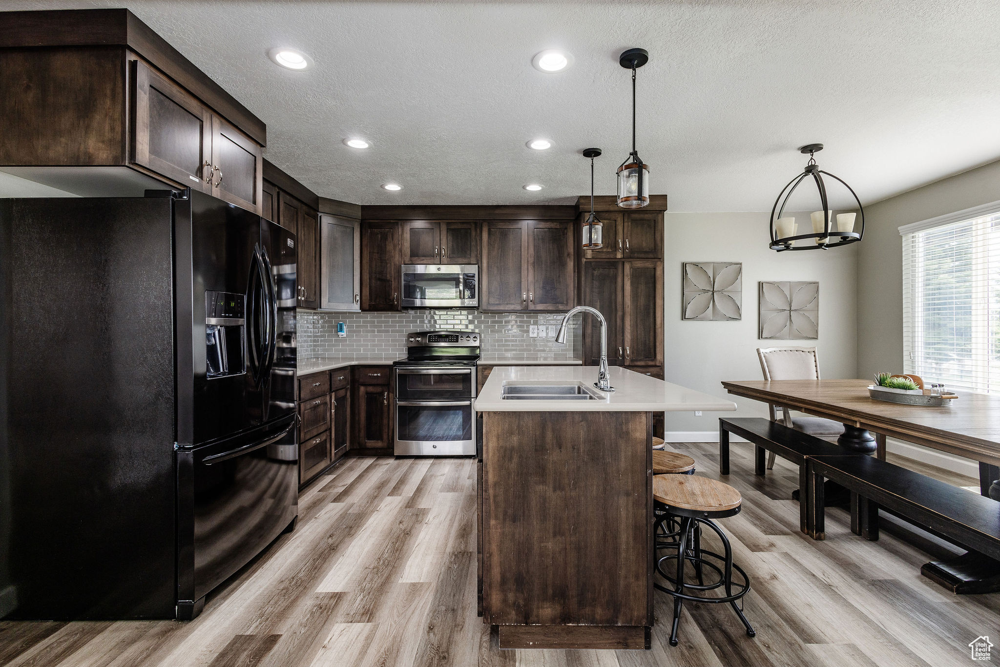 Kitchen featuring light hardwood / wood-style flooring, tasteful backsplash, hanging light fixtures, a kitchen island with sink, and appliances with stainless steel finishes