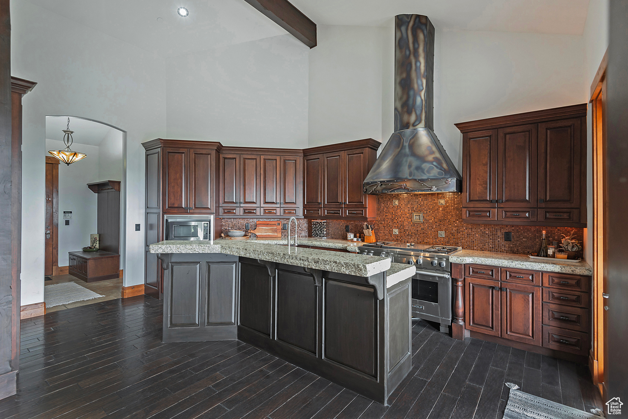 Kitchen featuring an island with sink, wall chimney exhaust hood, stainless steel appliances, and high vaulted ceiling