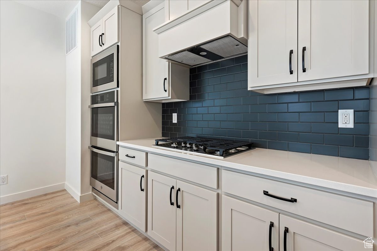 Kitchen featuring custom exhaust hood, stainless steel appliances, light wood-type flooring, backsplash, and white cabinets