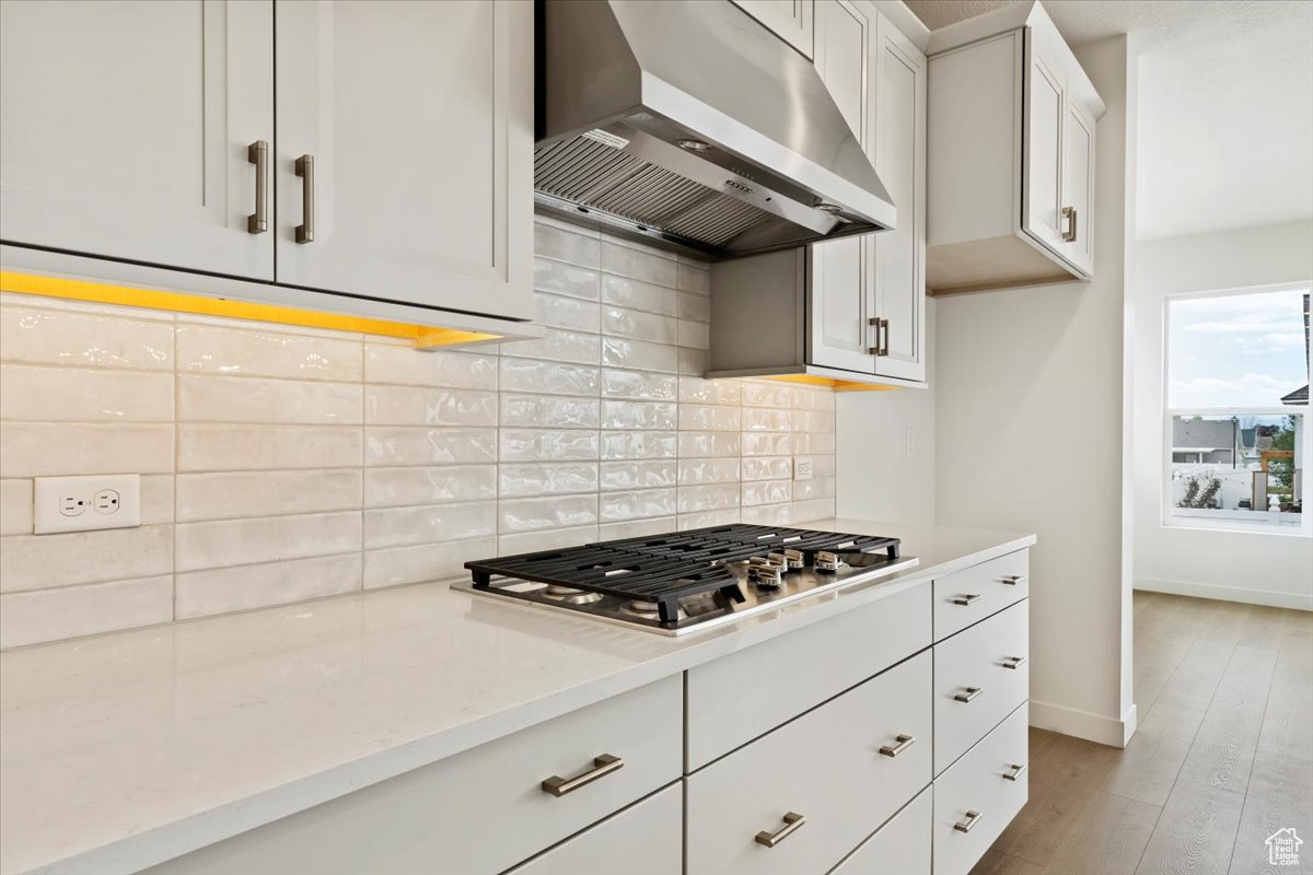 Kitchen featuring white cabinets, stainless steel gas stovetop, wall chimney range hood, decorative backsplash, and light wood-type flooring