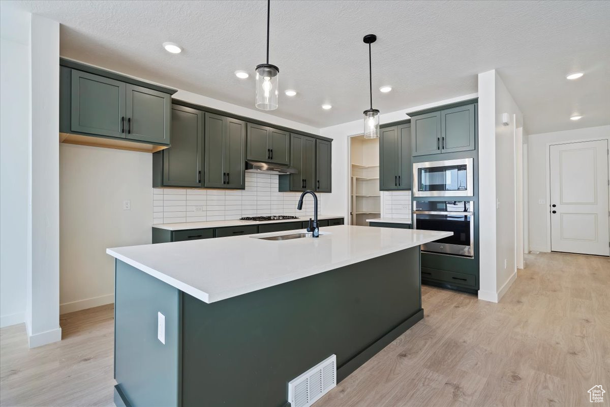 Kitchen featuring light wood-type flooring, stainless steel appliances, decorative backsplash, pendant lighting, and a center island with sink