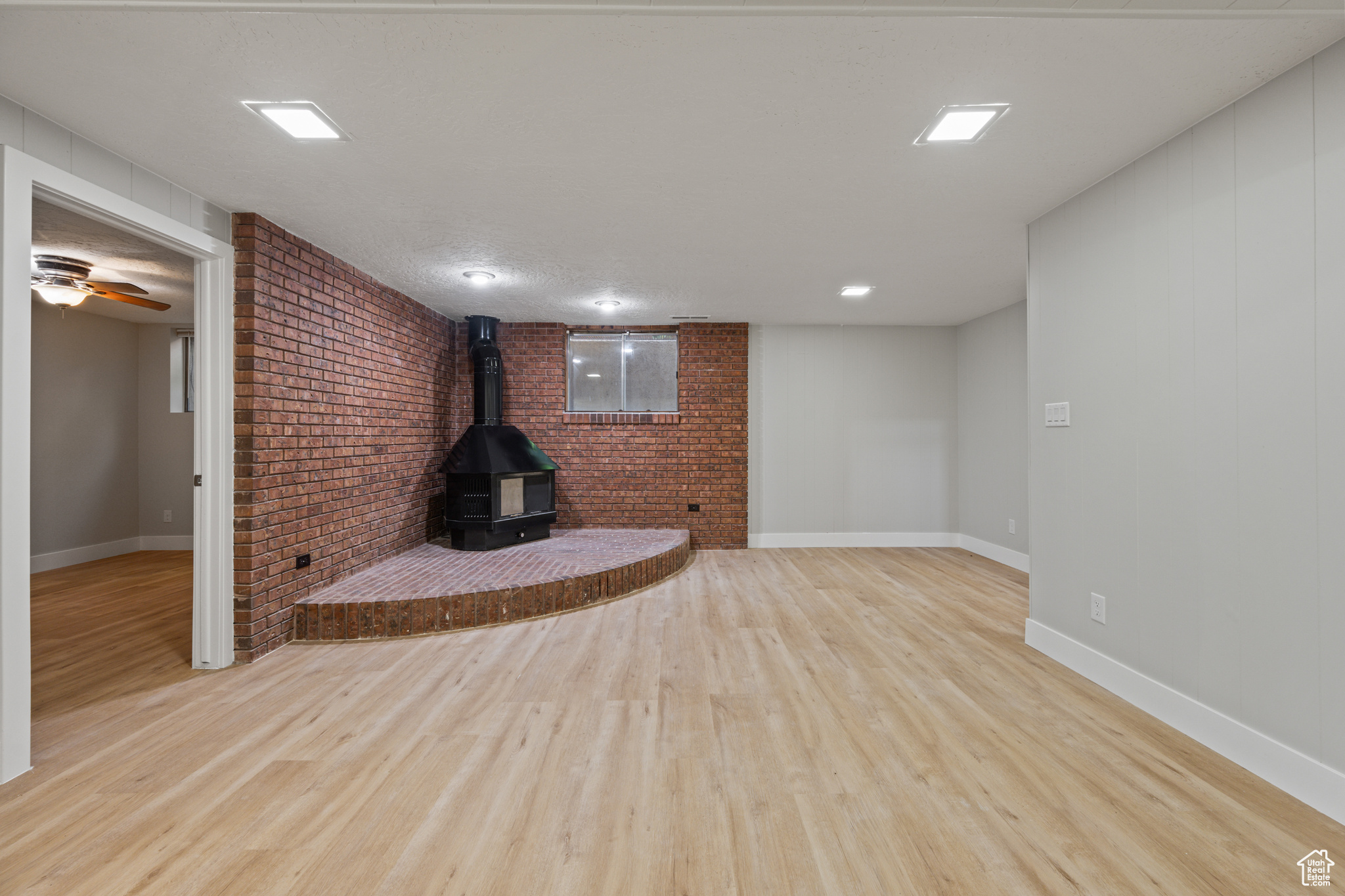 Unfurnished living room with ceiling fan, light wood-type flooring, a wood stove, and brick wall