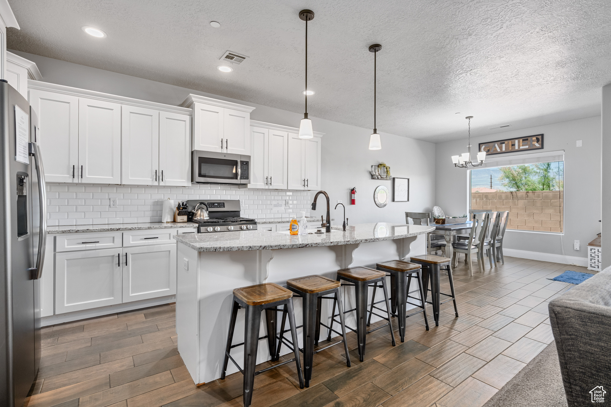 Kitchen with tasteful backsplash, an island with sink, stainless steel appliances, hanging light fixtures, and white cabinetry