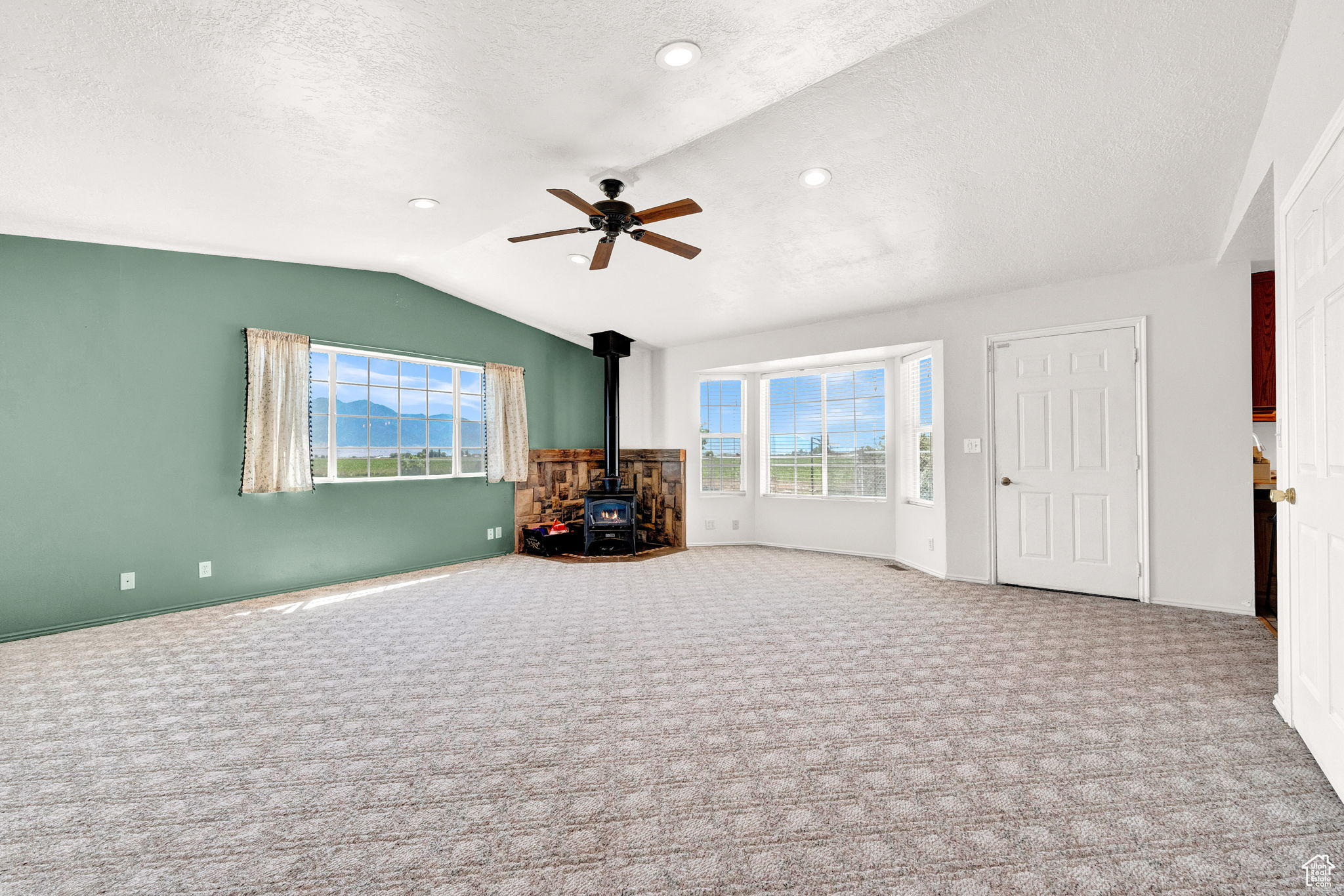 Unfurnished living room featuring ceiling fan, a wood stove, carpet floors, vaulted ceiling, and a textured ceiling