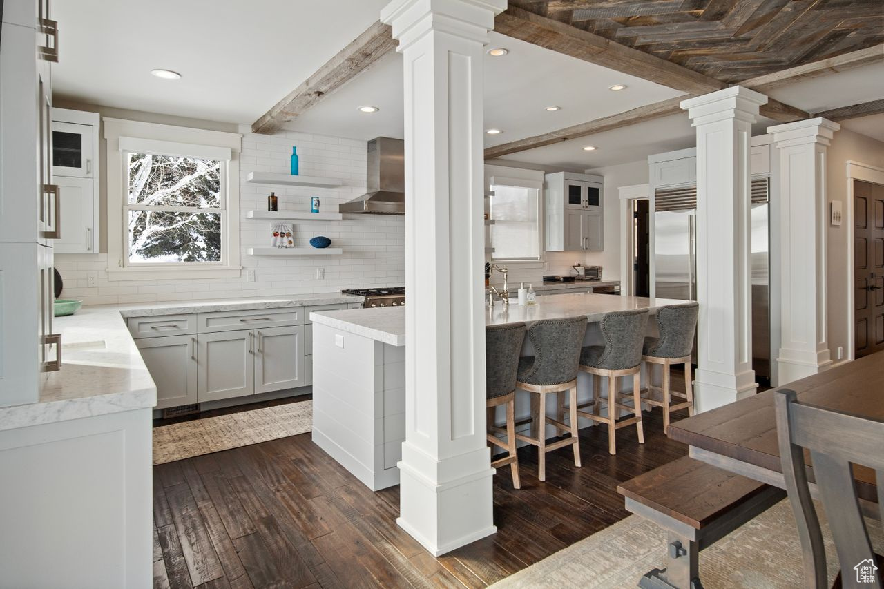 Kitchen with dark wood-type flooring, ornate columns, wall chimney exhaust hood, and tasteful backsplash